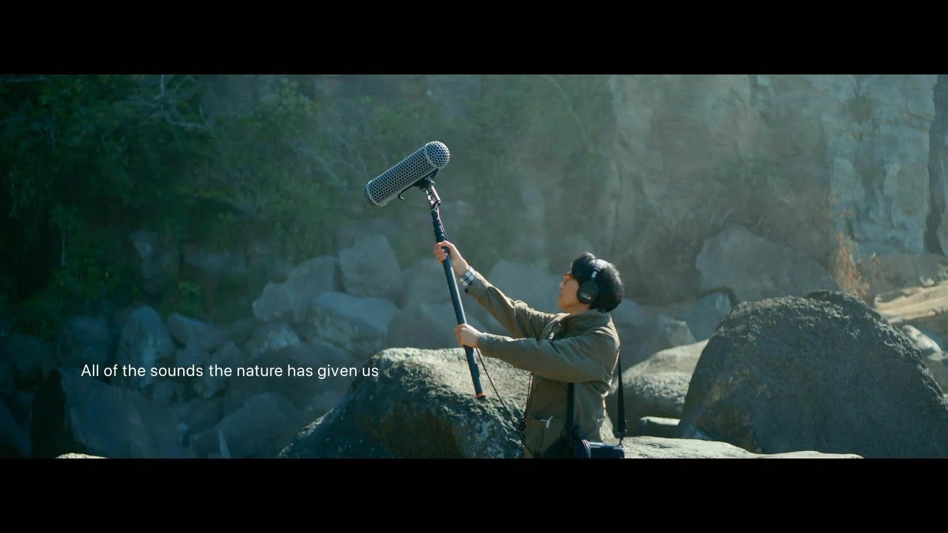 A person in a jacket and headphones holding a boom microphone in a rocky outdoor setting with trees, with a subtitle reading 'All of the sounds the nature has given us'.