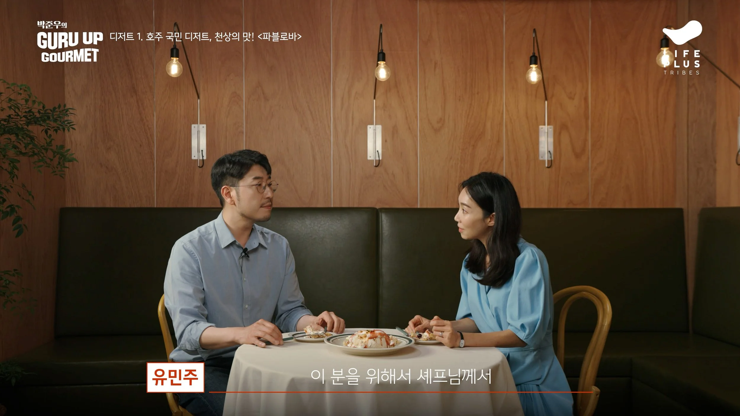 A man and woman sit across from each other at a table in a restaurant, having a conversation. The table has plates of food and a pizza. They are in a cozy, wooden-paneled setting with hanging light bulbs.