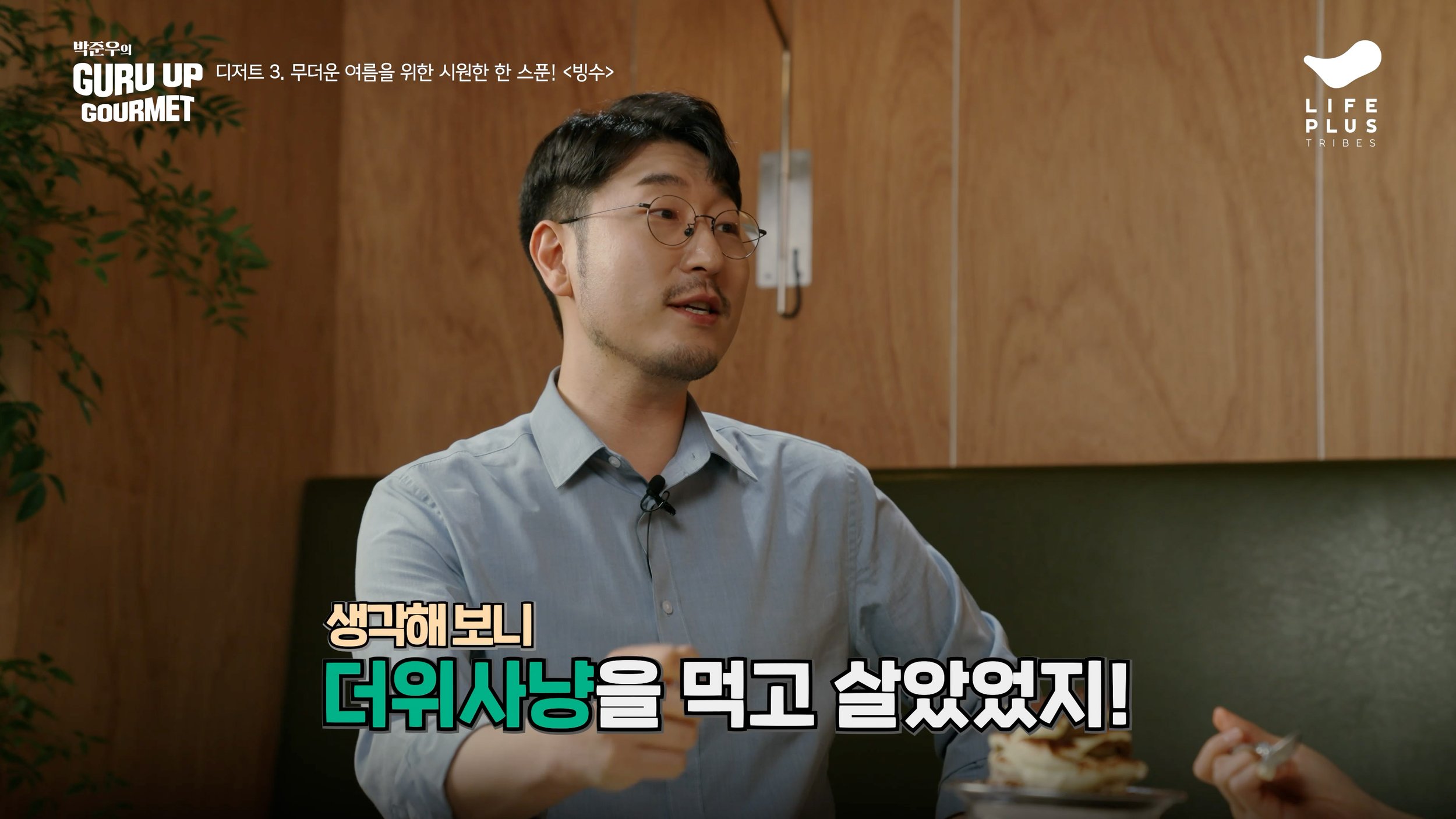 A man with glasses and a light blue shirt sitting at a table in a room with wooden walls, speaking with subtitles in Korean at the bottom.