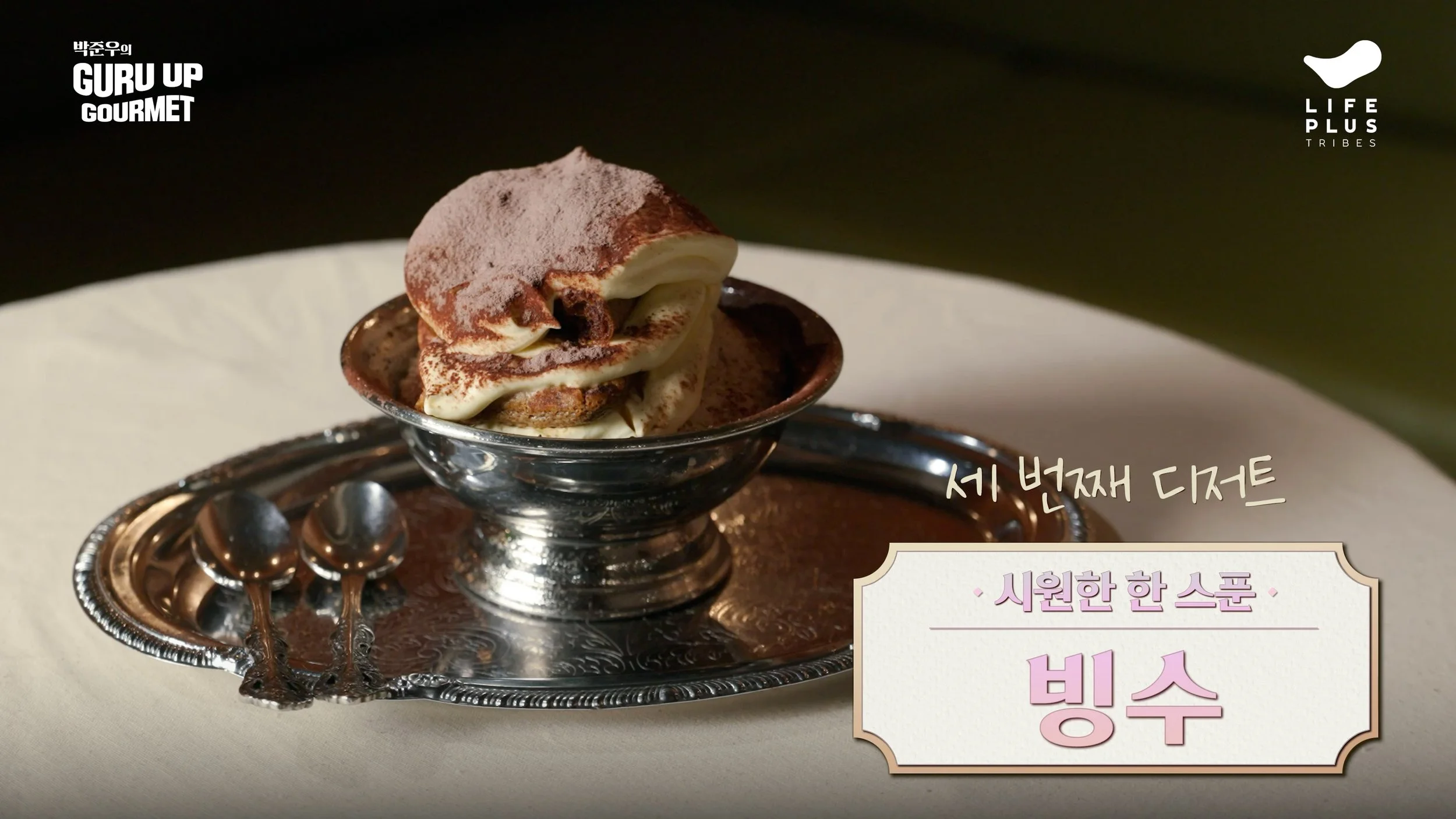 A small glass bowl of tiramisu on a decorative silver tray, with a spoon beside it, on a cream-colored tablecloth.