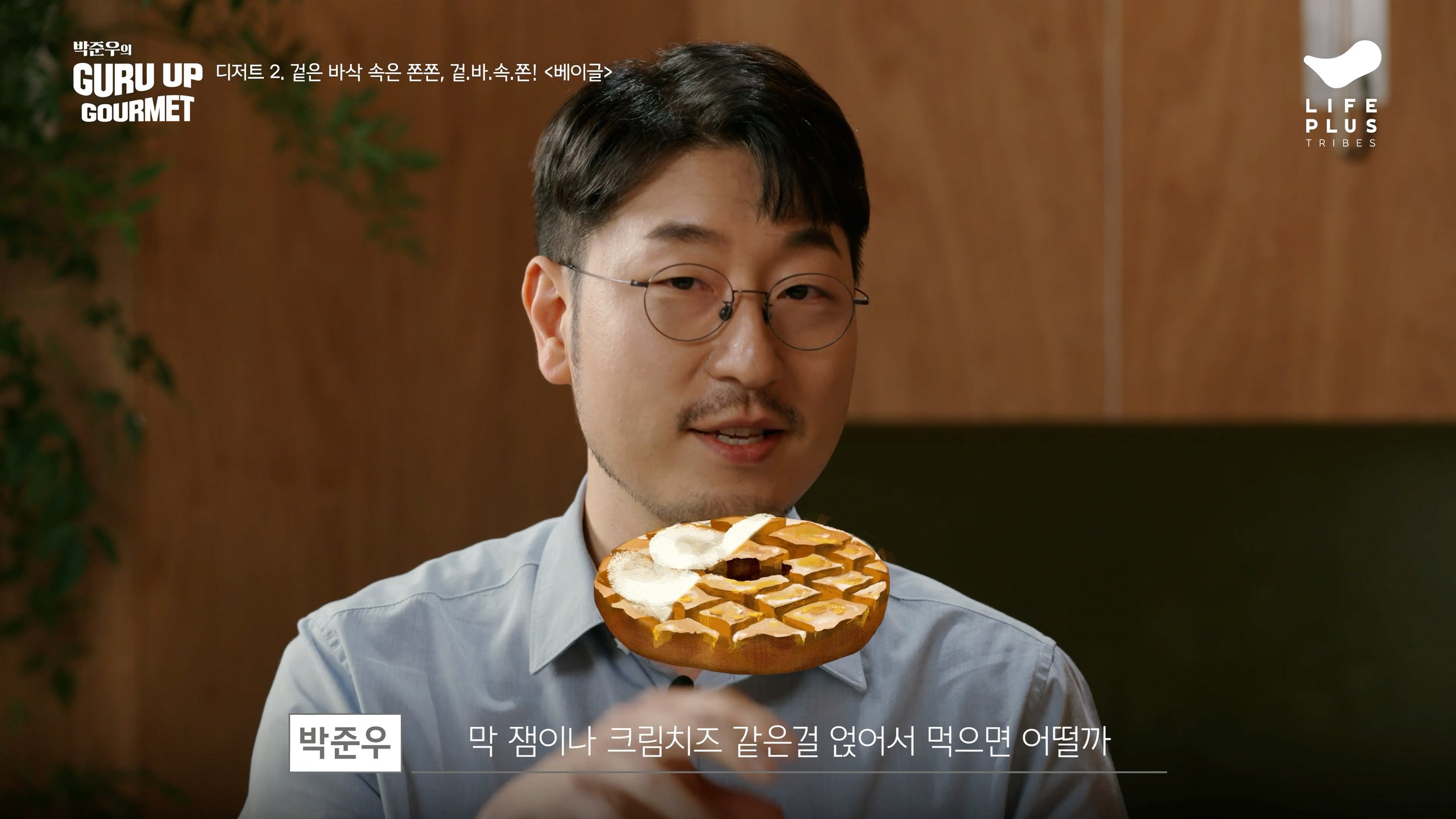 A man with glasses and a gray shirt is holding a plate with a waffle topped with whipped cream and caramel sauce. There is Korean text on the image, and a wooden background with plants on the side.