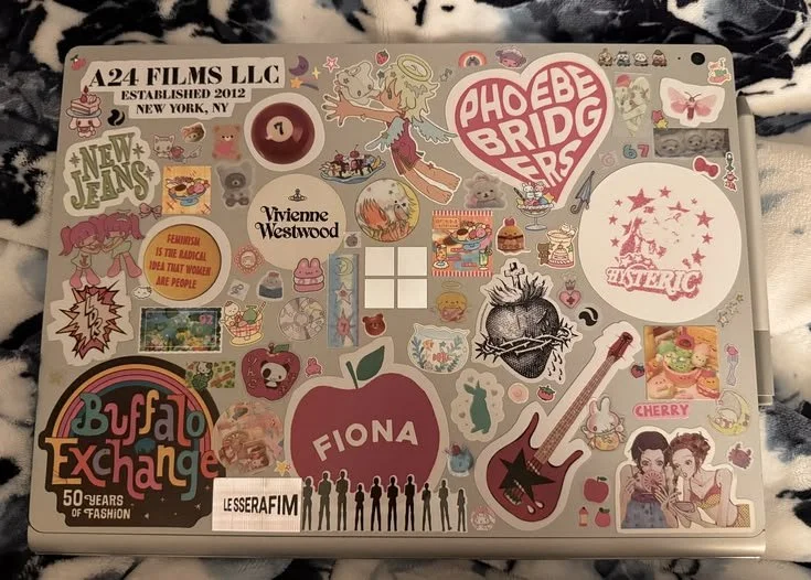 Laptop covered in various colorful stickers, including a large pink and white heart with "PHOEBE BRIDGE" text, a red apple with "FIONA" written inside, a rainbow with "Buffalo Exchange" text, a guitar, a portrait of two women, and many other small decorative stickers.