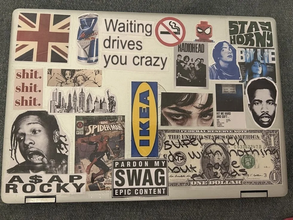 A laptop covered with various stickers including a British flag, a cup of tea, a no-smoking sign, radiohead, stay humble, a no-bath sticker, a Spider-Man comic cover, a one-dollar bill with scribbles, a large IKEA logo, a picture of a woman with dark eyes, a portrait of a man, and several text stickers such as "shit. shit. shit.", "A $ P R O C K Y", and "Pardon my swag epic content".