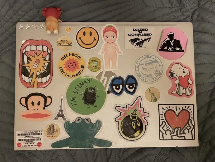 A laptop covered with various colorful stickers, including a smiley face, a doll, a watermelon, a frog, a heart, a monkey face, and other playful designs.