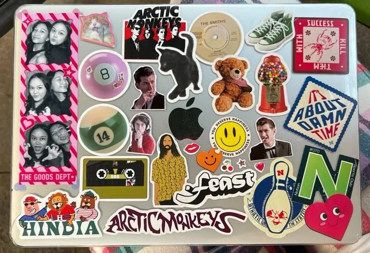 Various colorful stickers on a surface, including band logos, smiley face, a teddy bear, a gumball machine, 'About Damn Time' text, a pair of green shoes, and several other images and designs.