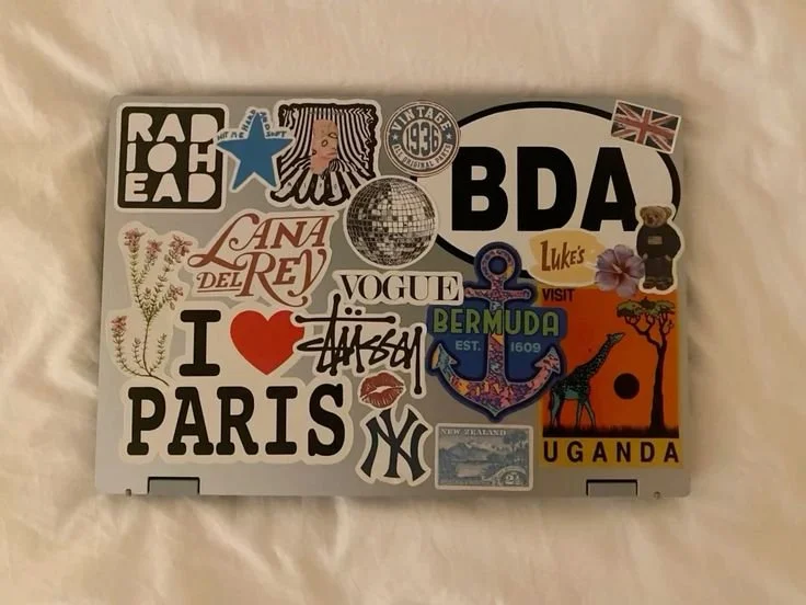 Laptop covered with assorted travel and fashion stickers, including references to Paris, Vogue, Bermuda, Uganda, and various symbols and logos.