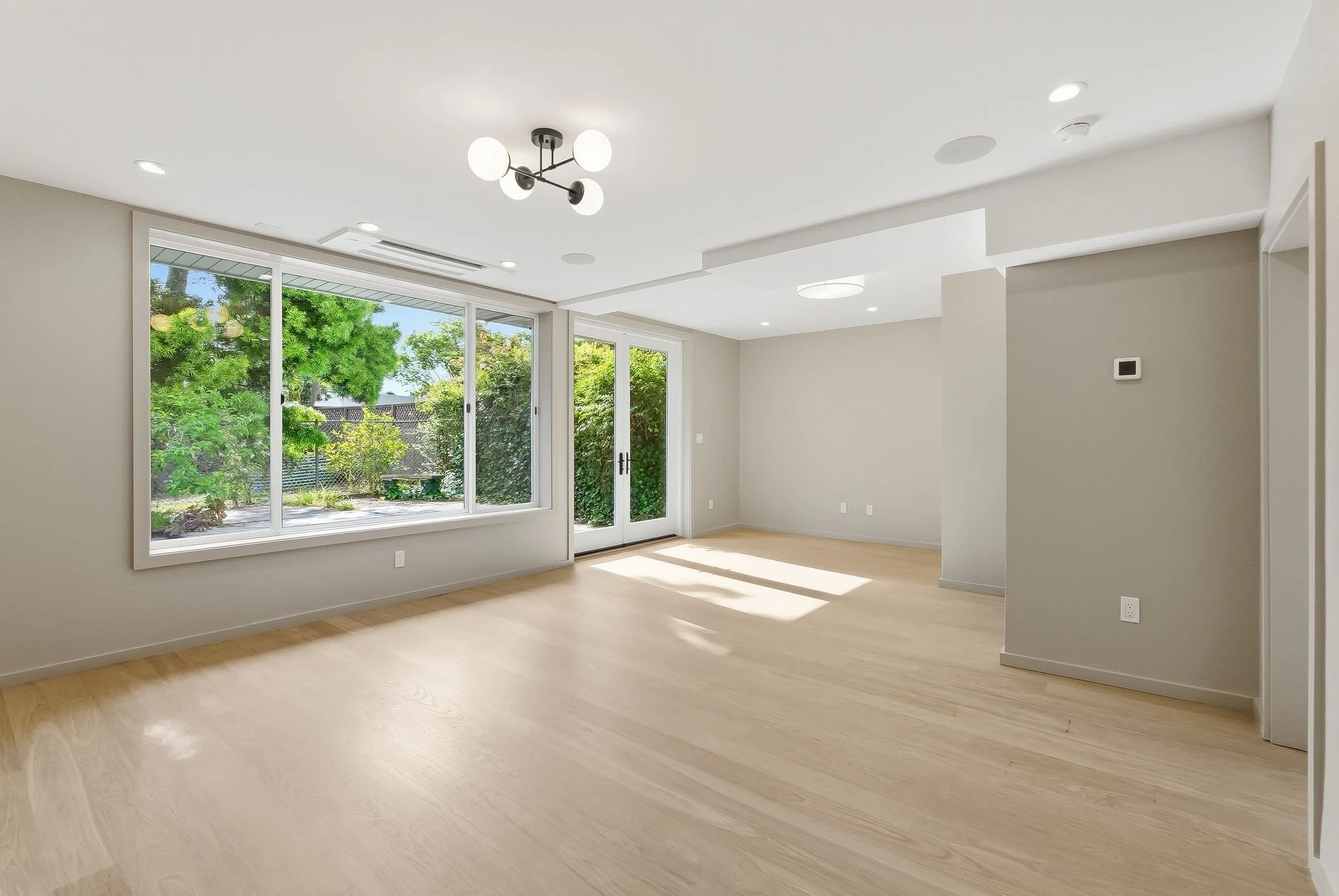Bright empty living room with large windows, light hardwood floors, and recessed lighting