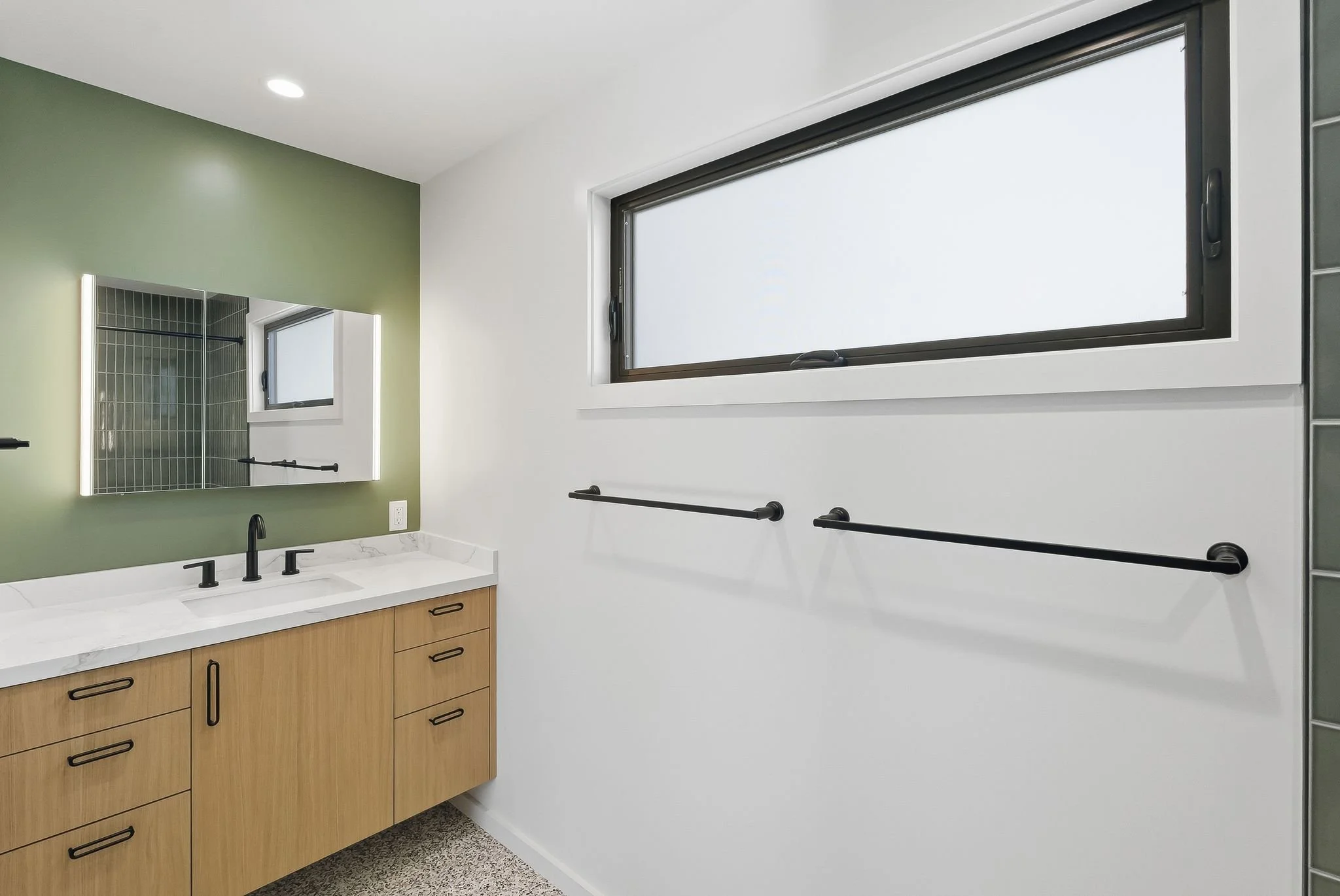 Long bathroom with double wood vanities, white countertops, green accent wall, and a large horizontal mirror
