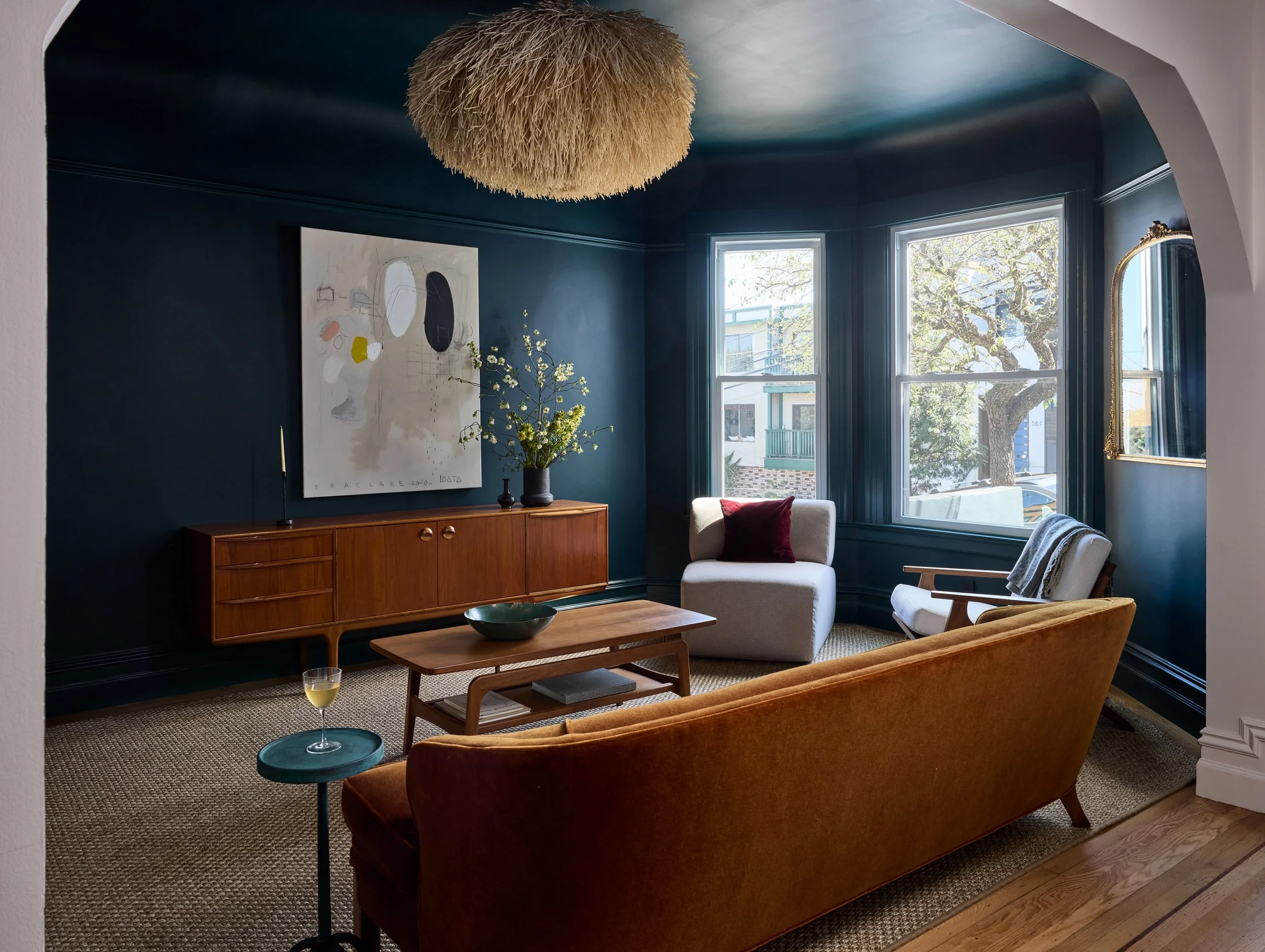 Noe Valley living and dining room with deep navy walls, a cognac leather sofa, mid-century wood credenza, and bay windows