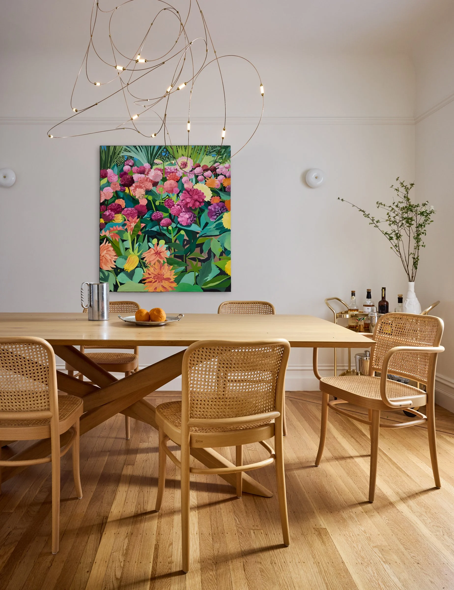 Dining room with a light oak table, woven cane chairs, colorful floral artwork, and a sculptural chandelier