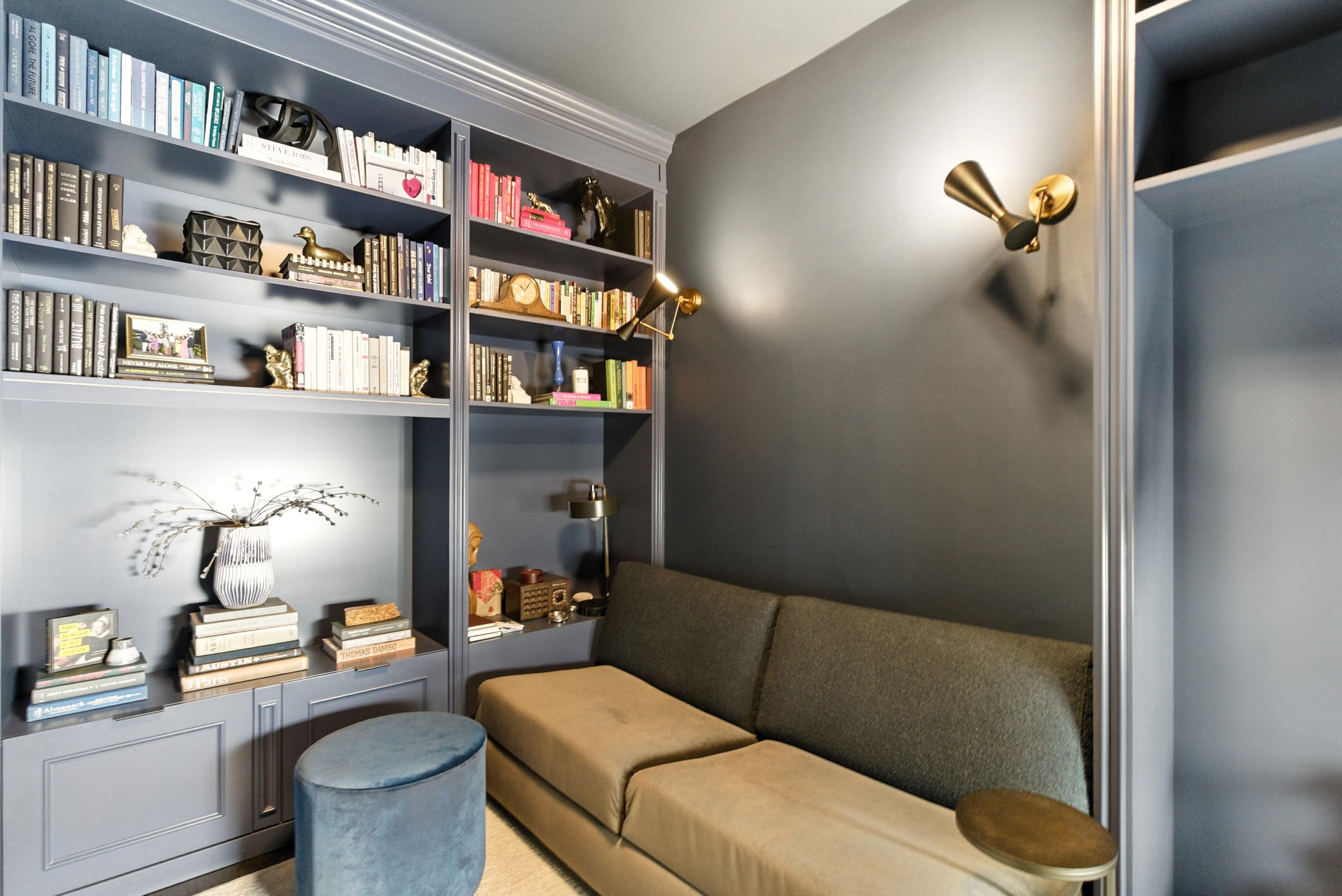 Cozy study with gray built-in bookshelves, an olive sofa, blue velvet ottoman, and brass sconce lighting