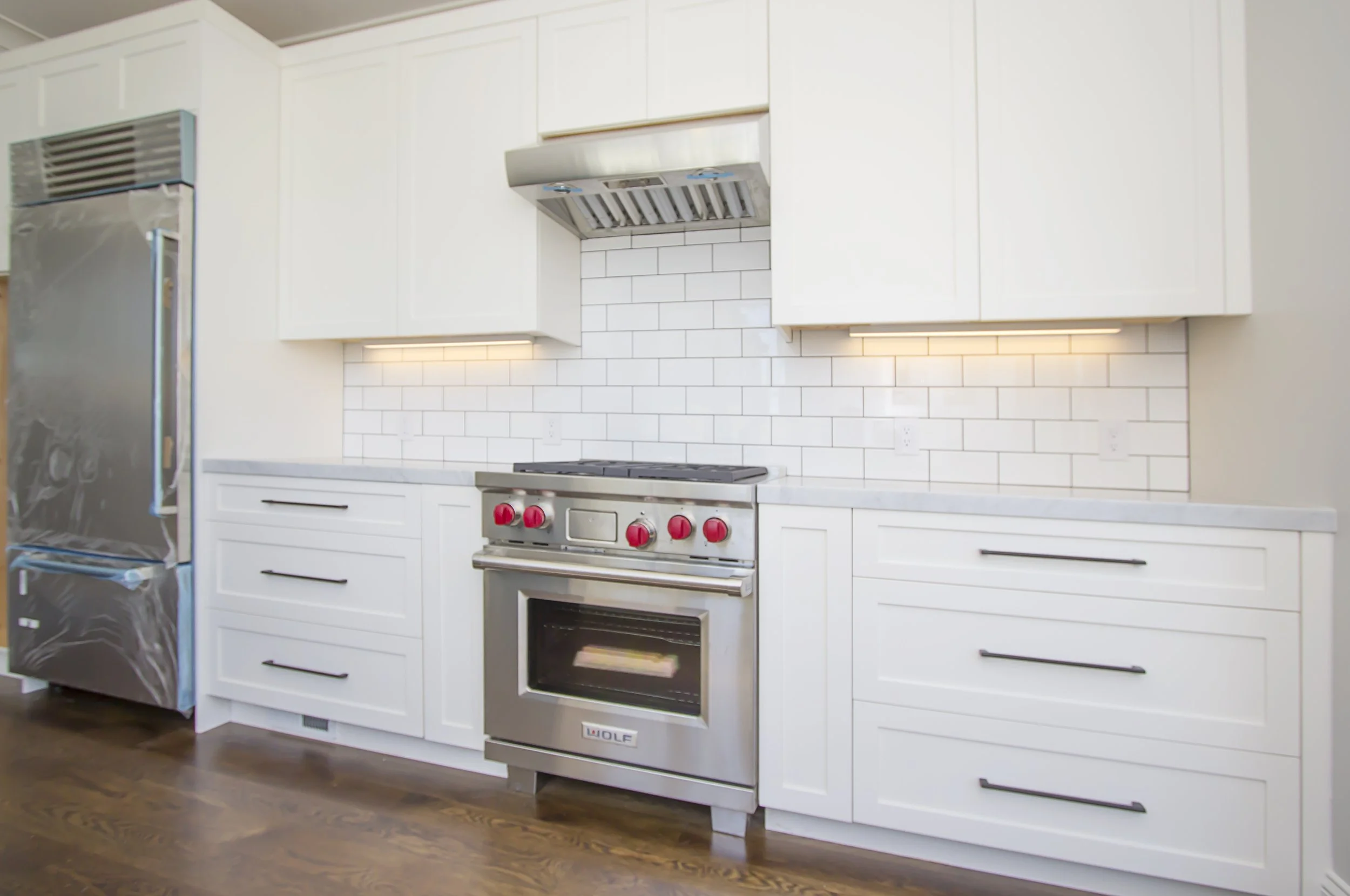 White kitchen with a stainless range and hood, a stainless refrigerator, a subway tile backsplash, and hardwood floors