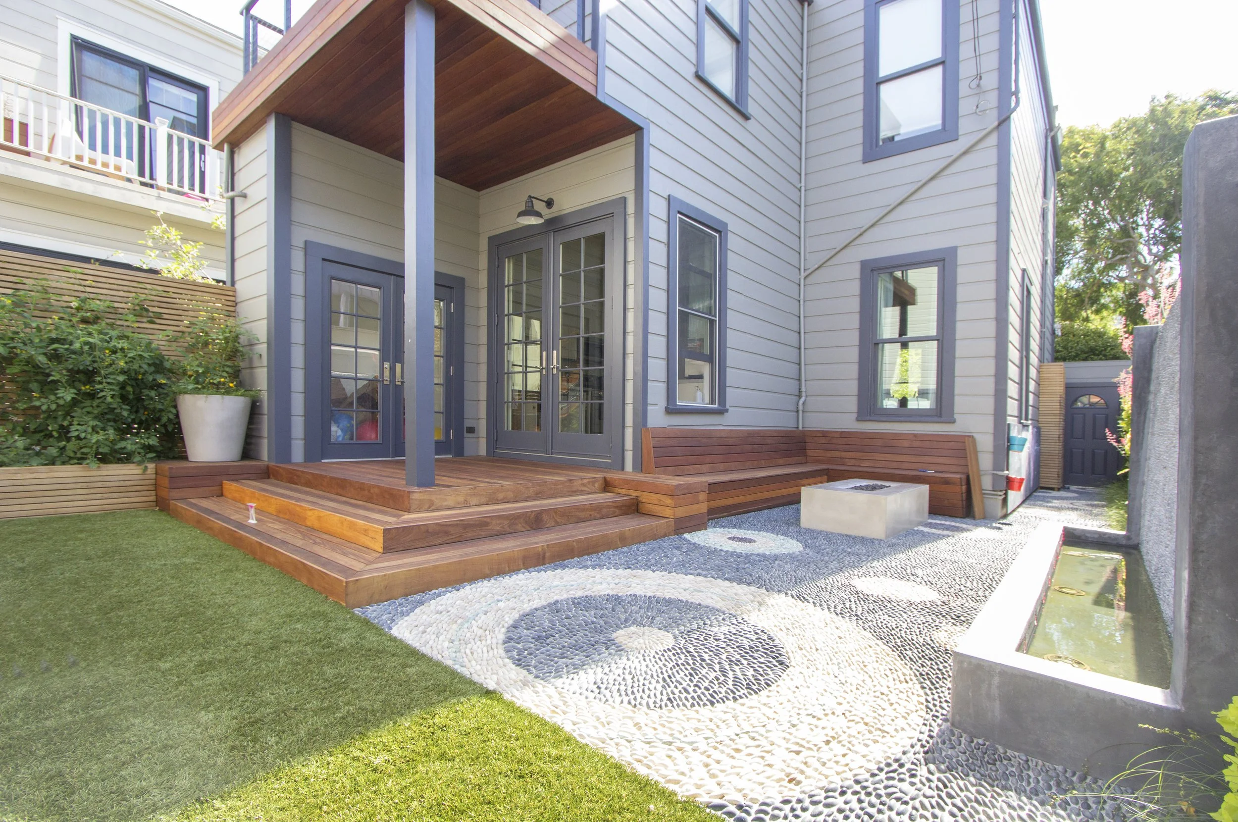 Rear entry deck with wood steps and posts leading to glass doors, beside a turf lawn and pebble border