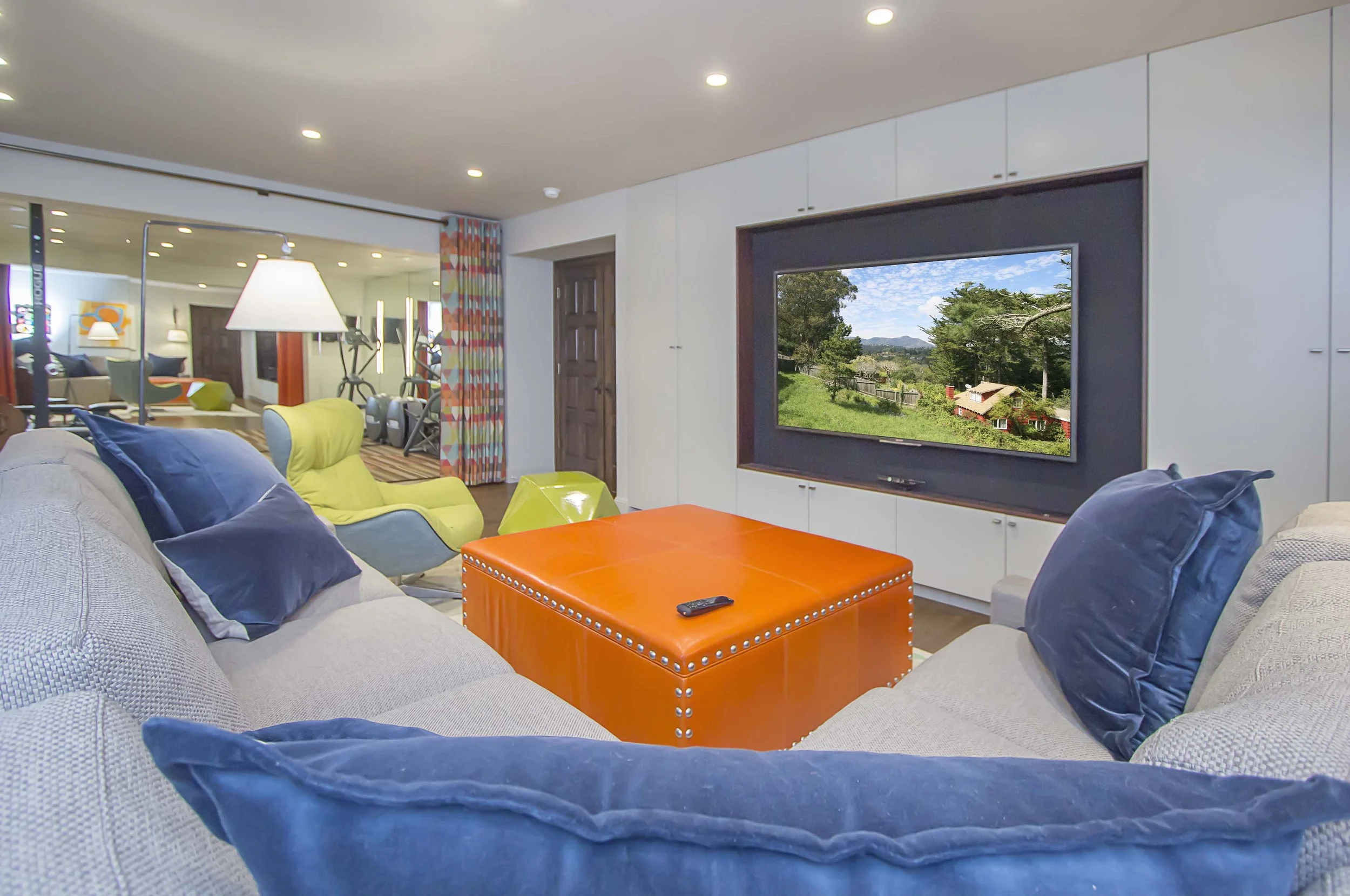 Media room with a large wall-mounted TV, an orange leather ottoman, and navy pillows on a sectional sofa
