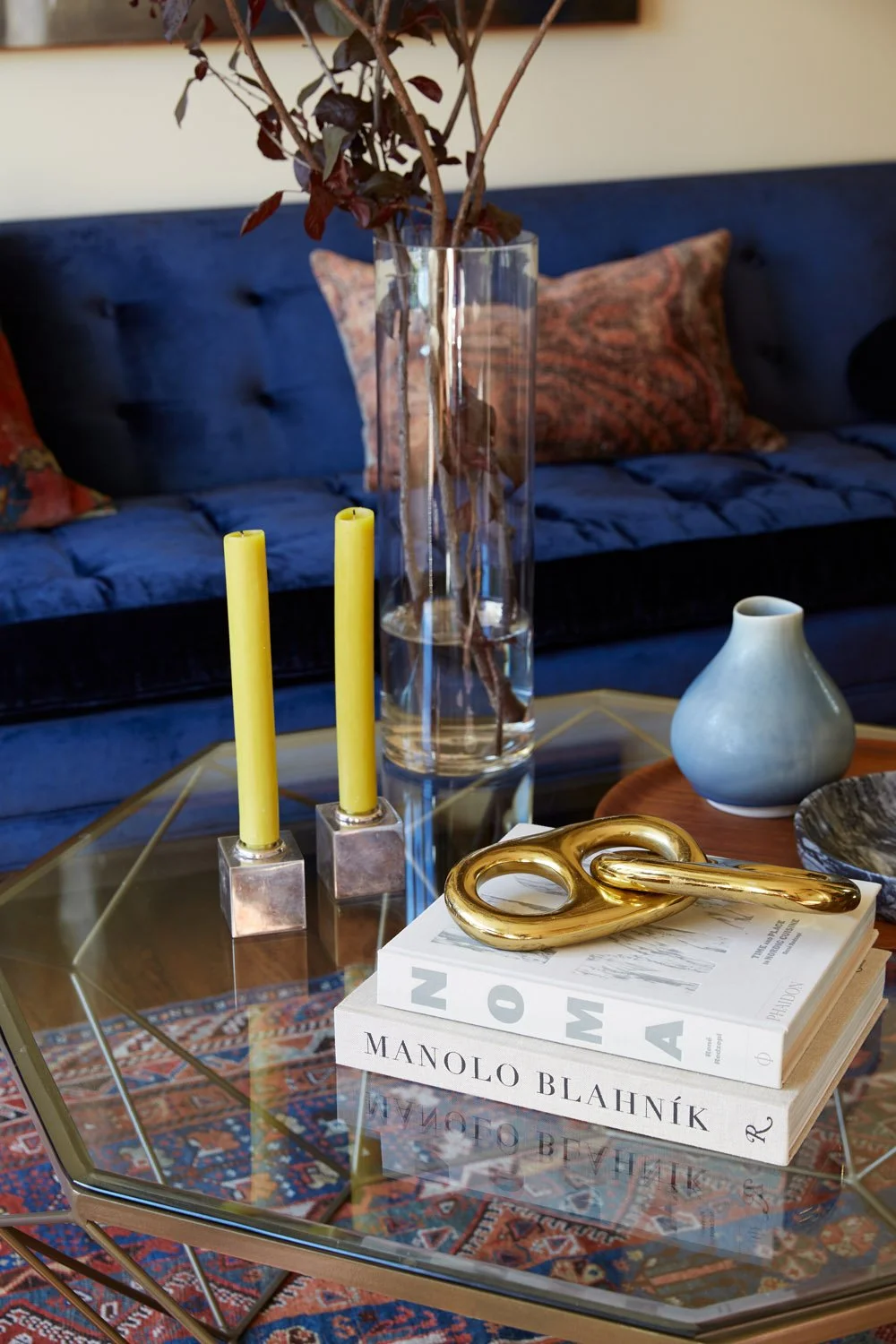 Glass coffee table styled with candlesticks, books, and decor in front of a blue tufted sofa