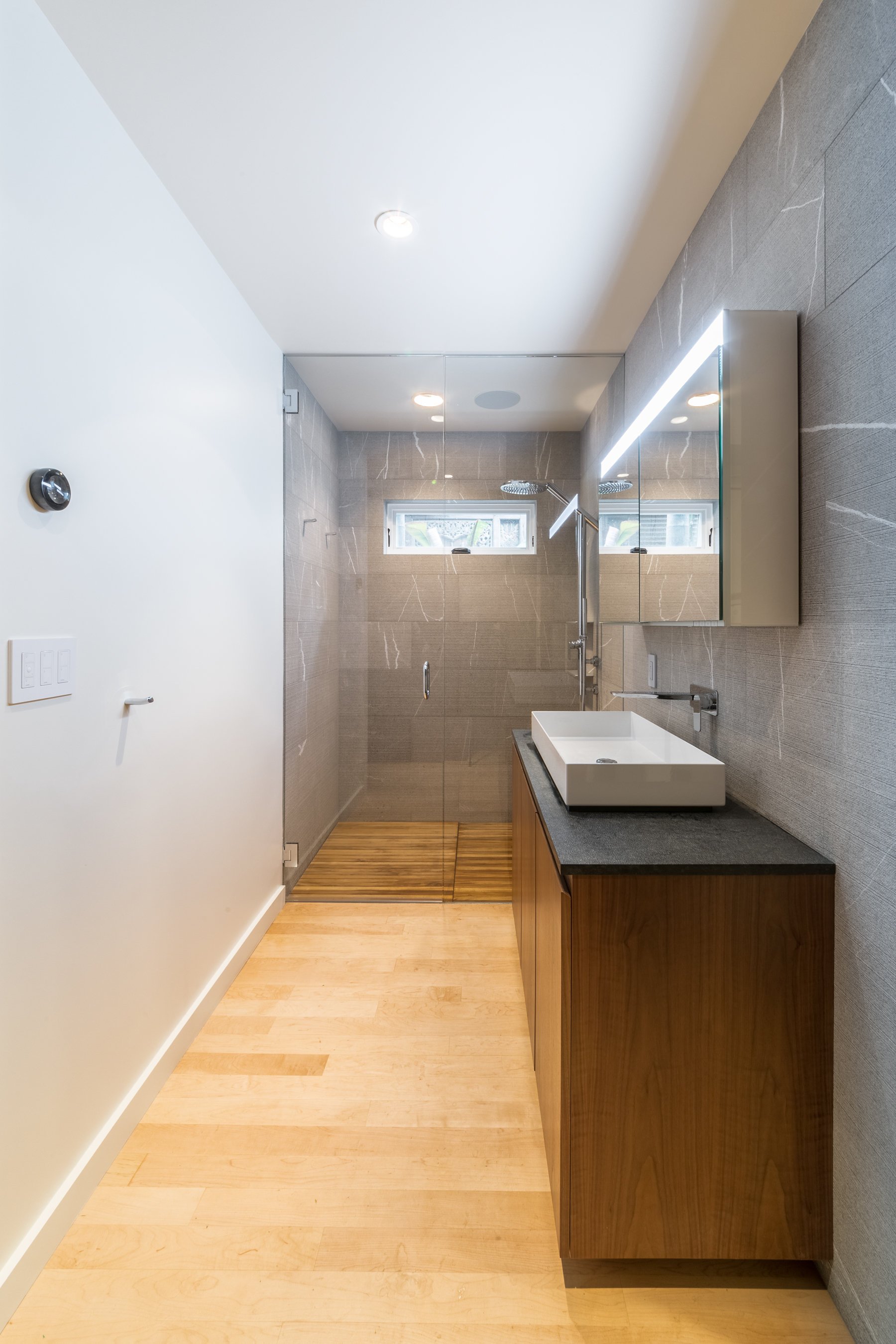 Long view of the grey stone-tiled bathroom, with maple flooring running into the curbless glass shower