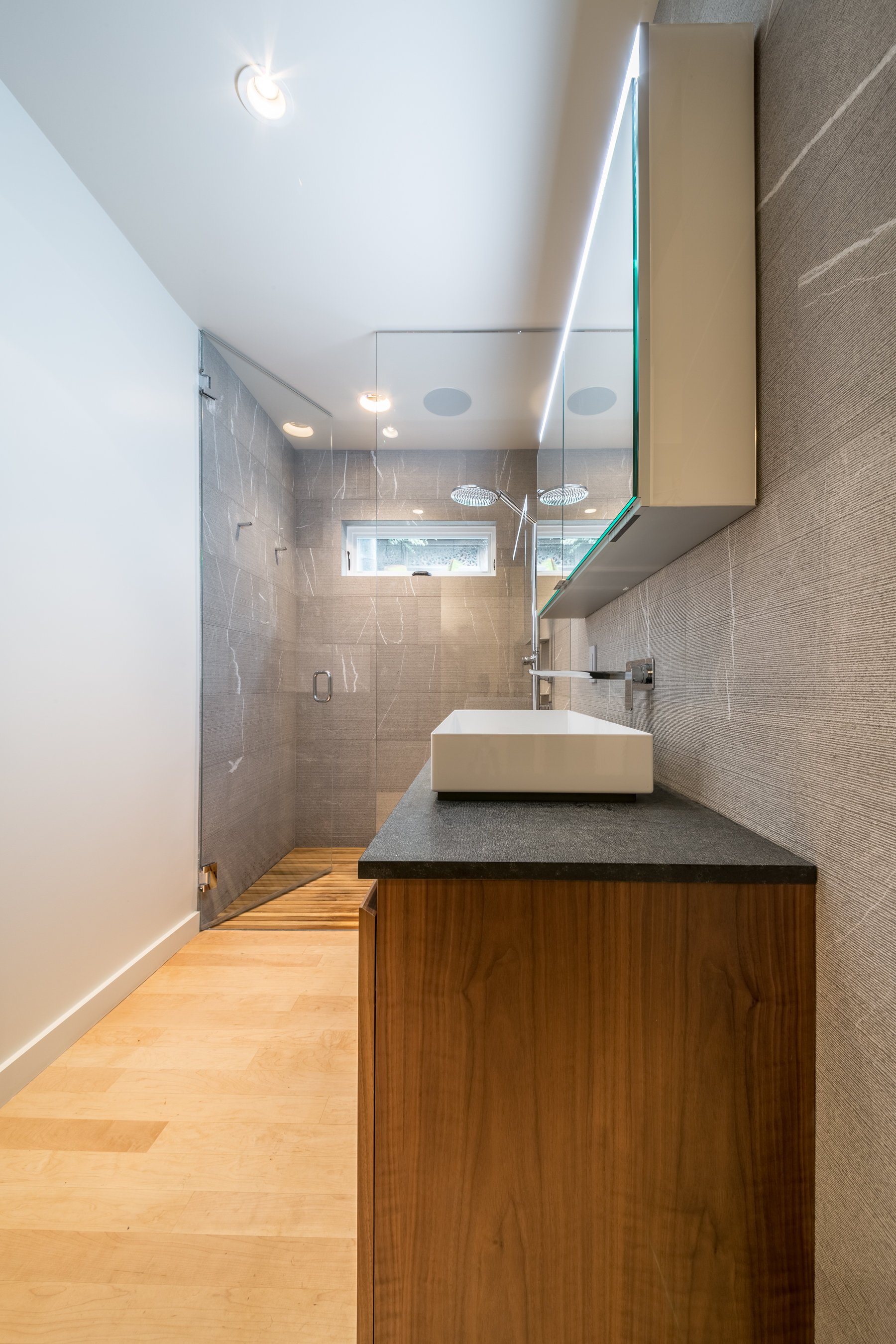 Walnut vanity and vessel sink along the grey stone wall, looking toward the glass shower