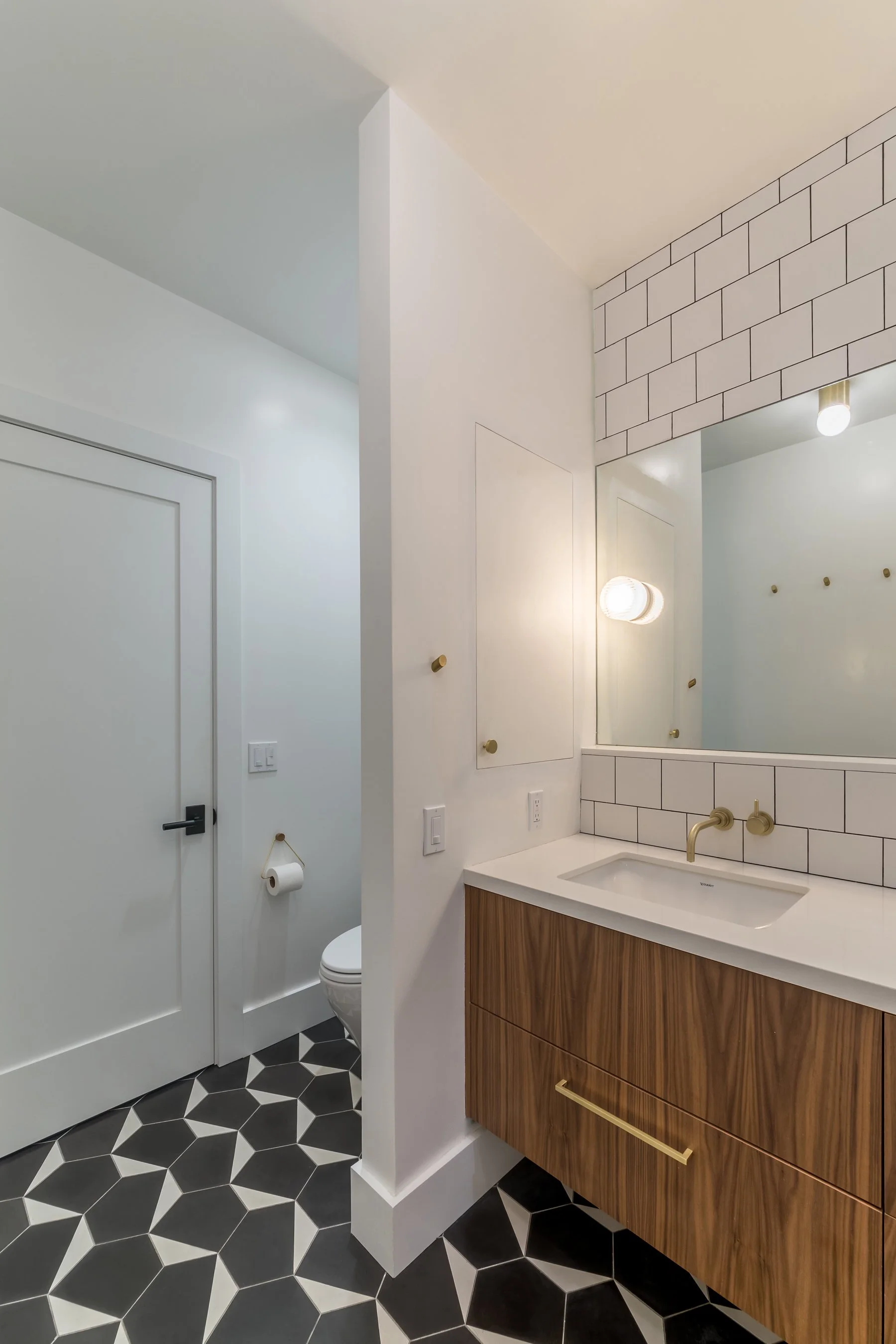White square-tiled bathroom with a walnut vanity, brass fixtures and a black-and-white geometric cement tile floor