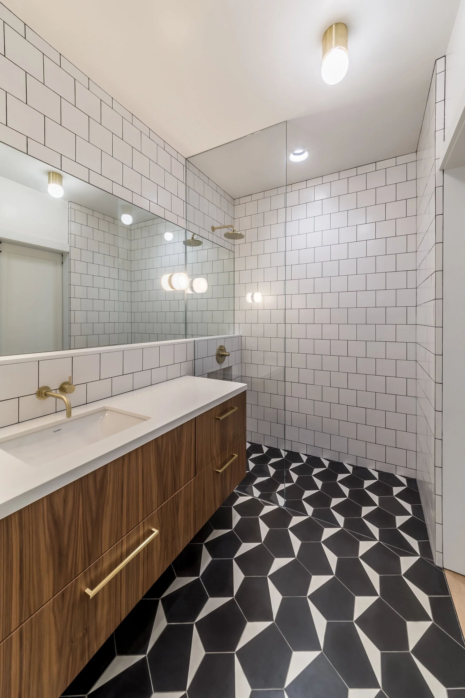 Floating walnut vanity with brass hardware facing the glass-walled shower, white square tile throughout