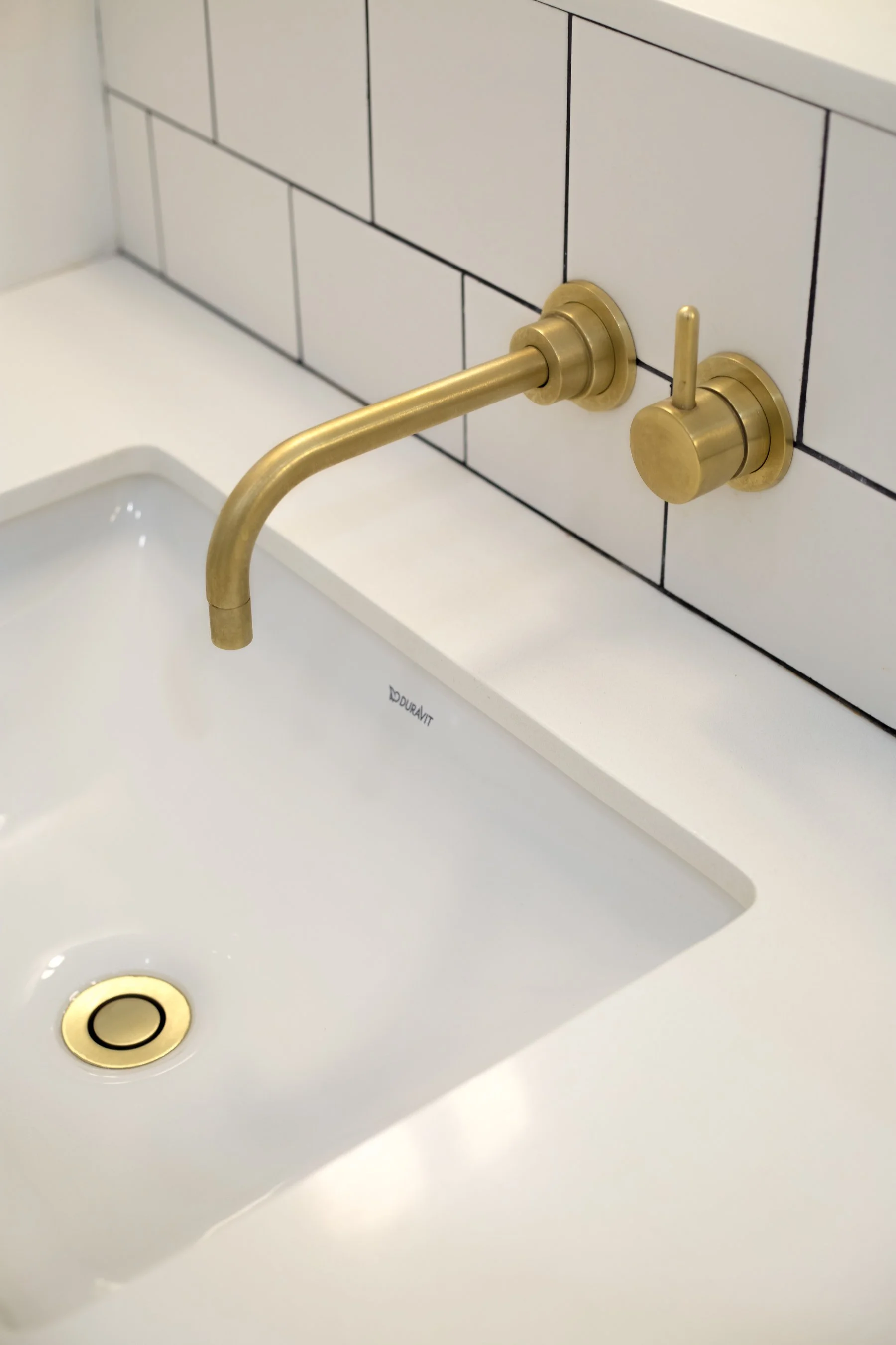 Brushed brass wall-mounted faucet over the undermount basin against white square tile