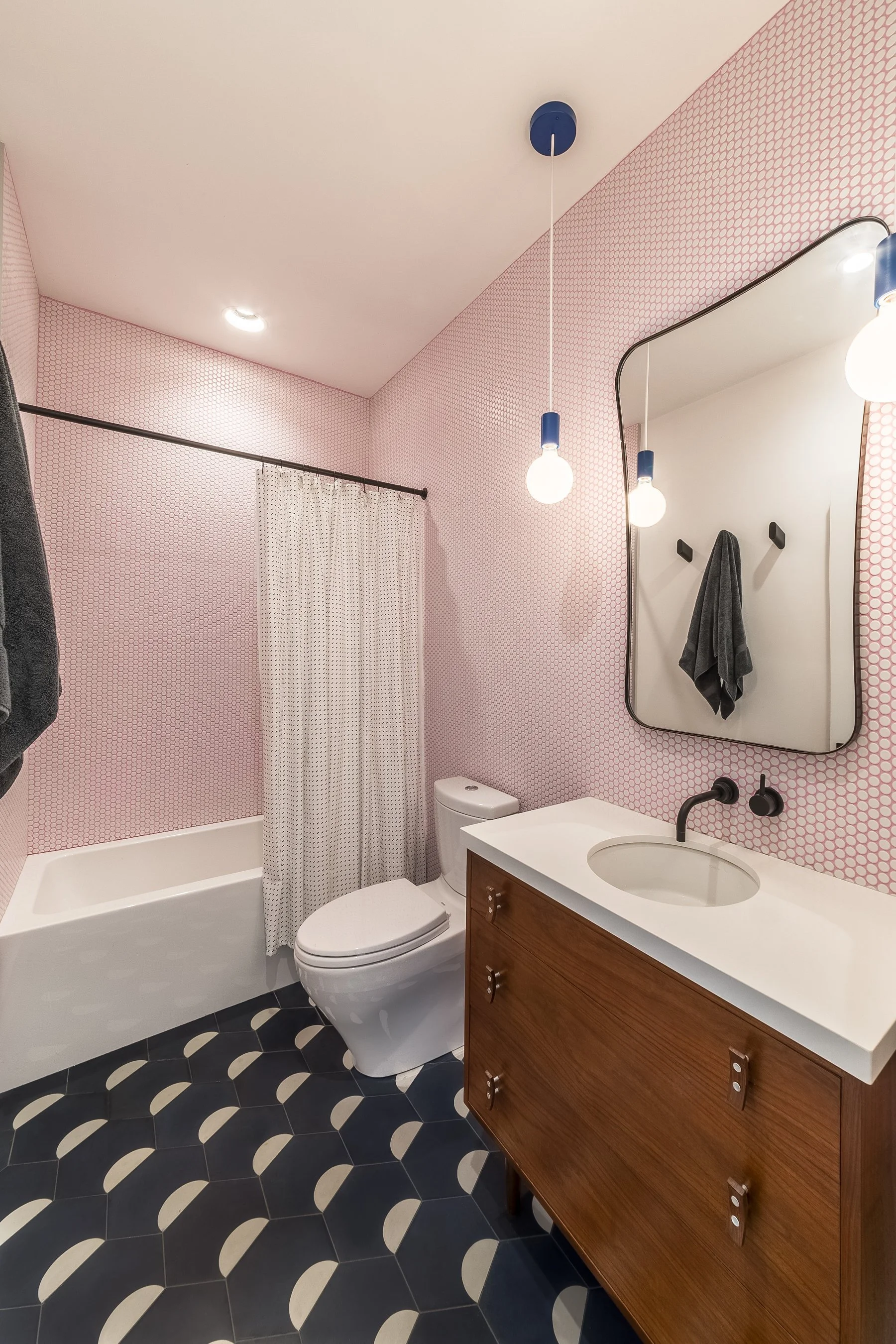Pink penny-tiled bathroom with a walnut vanity, matte black fixtures, blue pendant lights and a navy-and-white tile floor