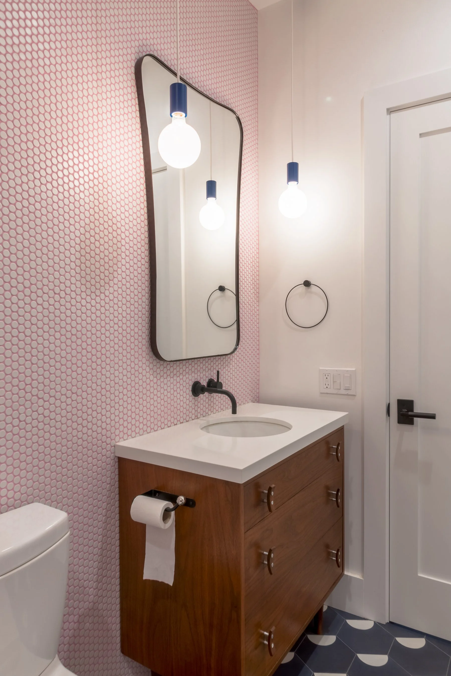 Arched black-framed mirror and blue pendant lights above the walnut vanity against pink penny tile