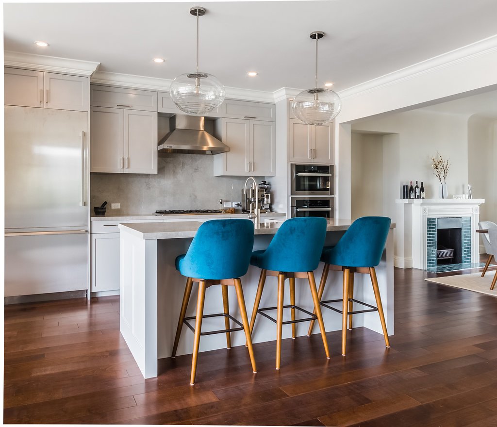 Open white kitchen with an island, blue bar stools, glass globe pendant lights, stainless hood, and dark hardwood floors