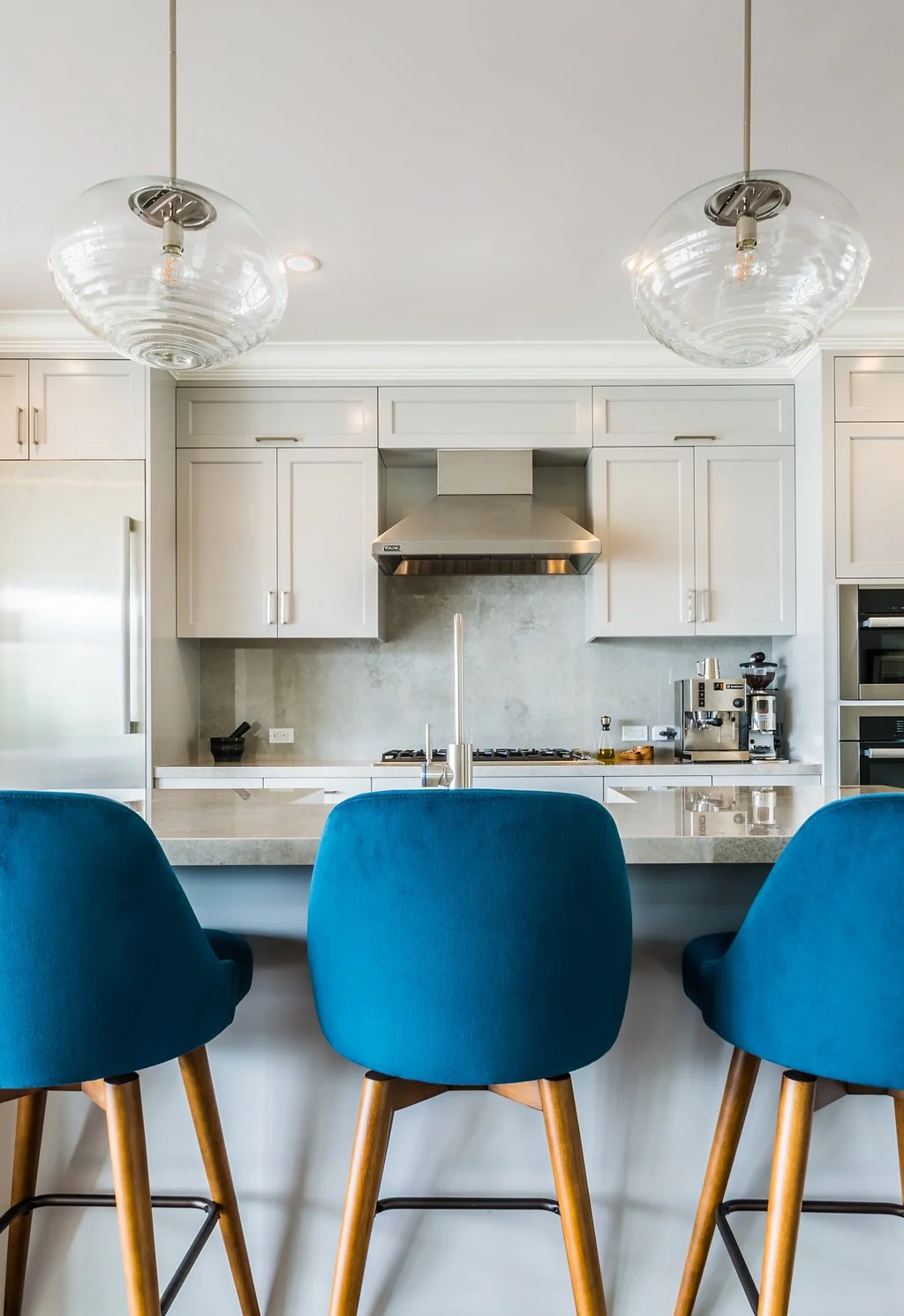 White kitchen island with blue bar stools, glass globe pendant lights, and a marble backsplash