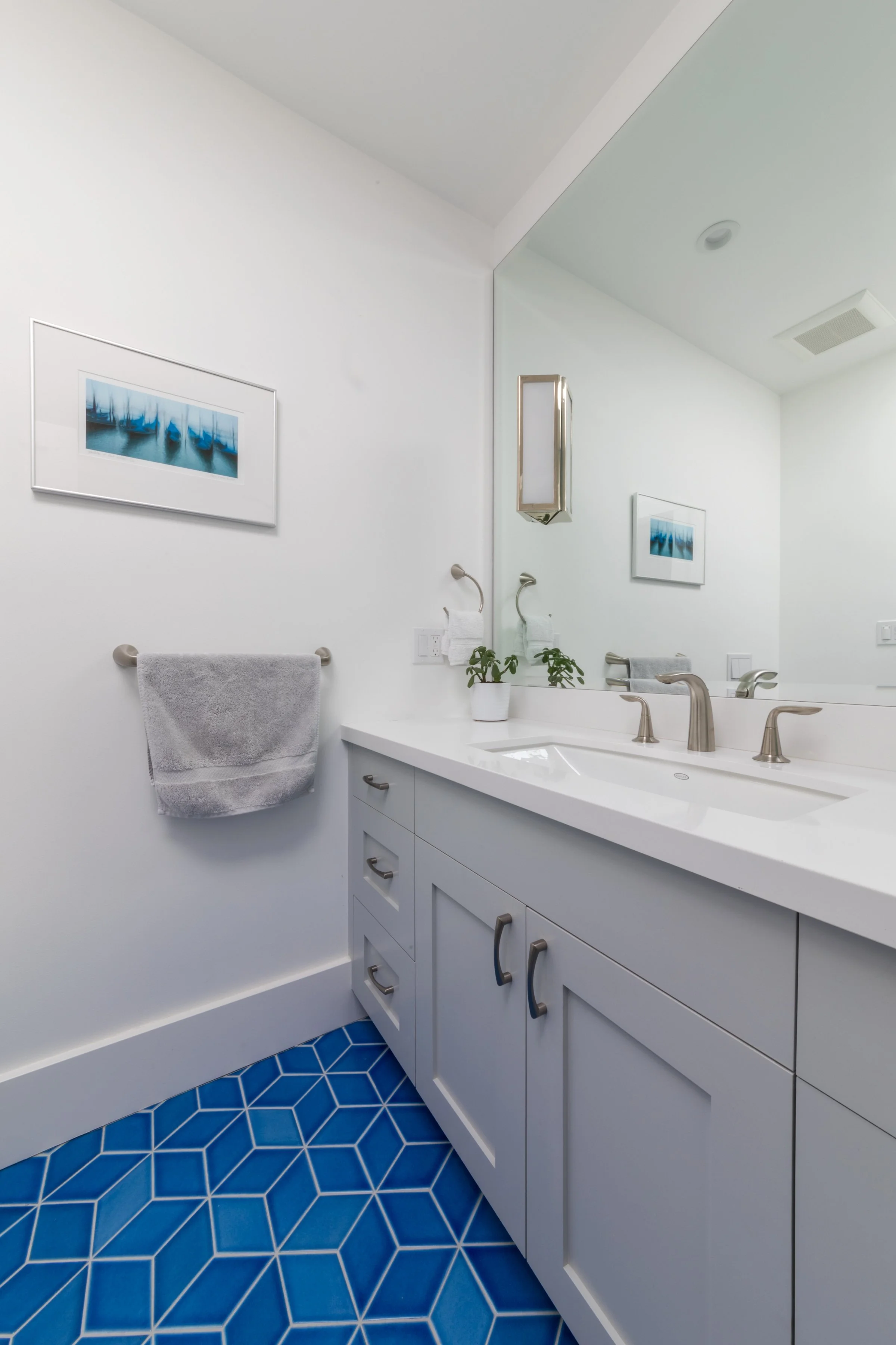 Double-vanity bathroom with gray cabinets, dual faucets, a wide mirror, coastal art, and blue geometric floor tile