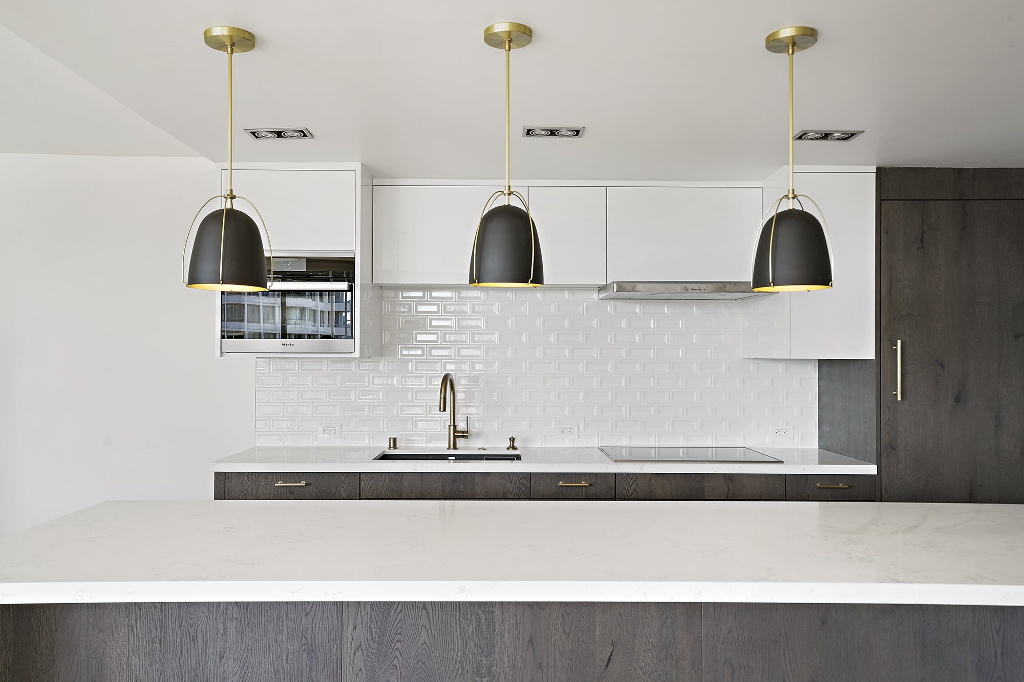 White kitchen with black dome pendant lights, dark wood cabinets, white beveled subway-tile backsplash, and a brass faucet