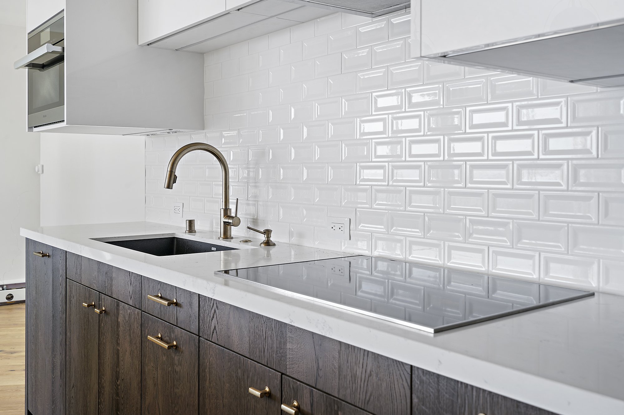 Kitchen counter with a dark wood base cabinet, white quartz countertop, cooktop, brass faucet, and beveled subway-tile backsplash