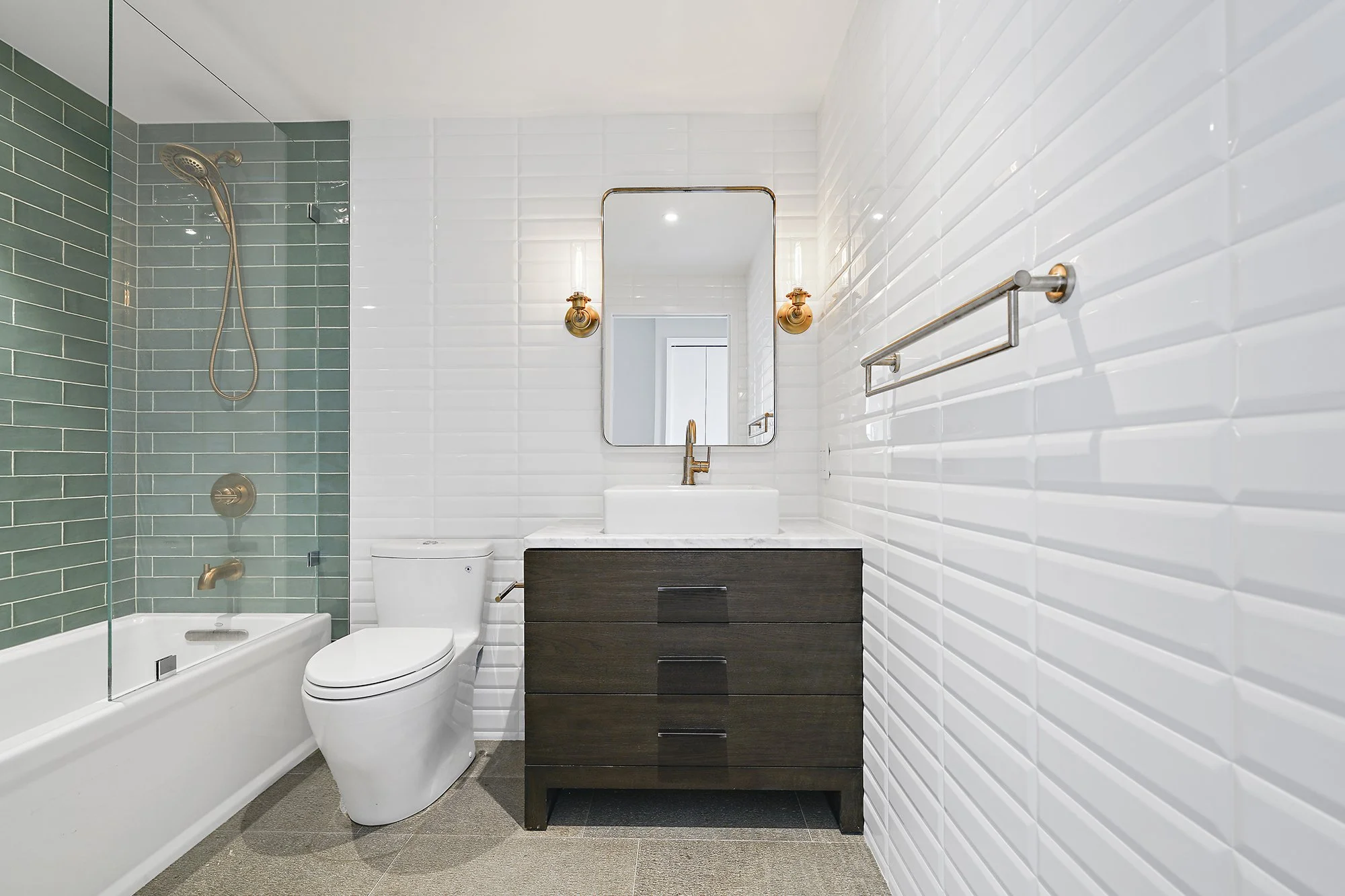 Bathroom with a white subway-tile wall, dark wood vanity, brass fixtures, a mirror, and a tub with a green-tiled surround
