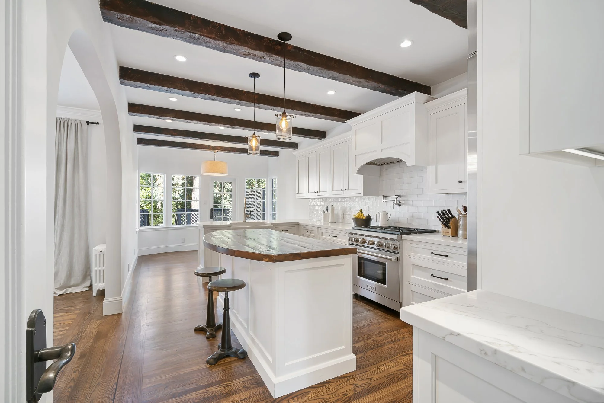 Open white kitchen with exposed dark wood ceiling beams, a wood-and-marble island, stainless range, and hardwood floors
