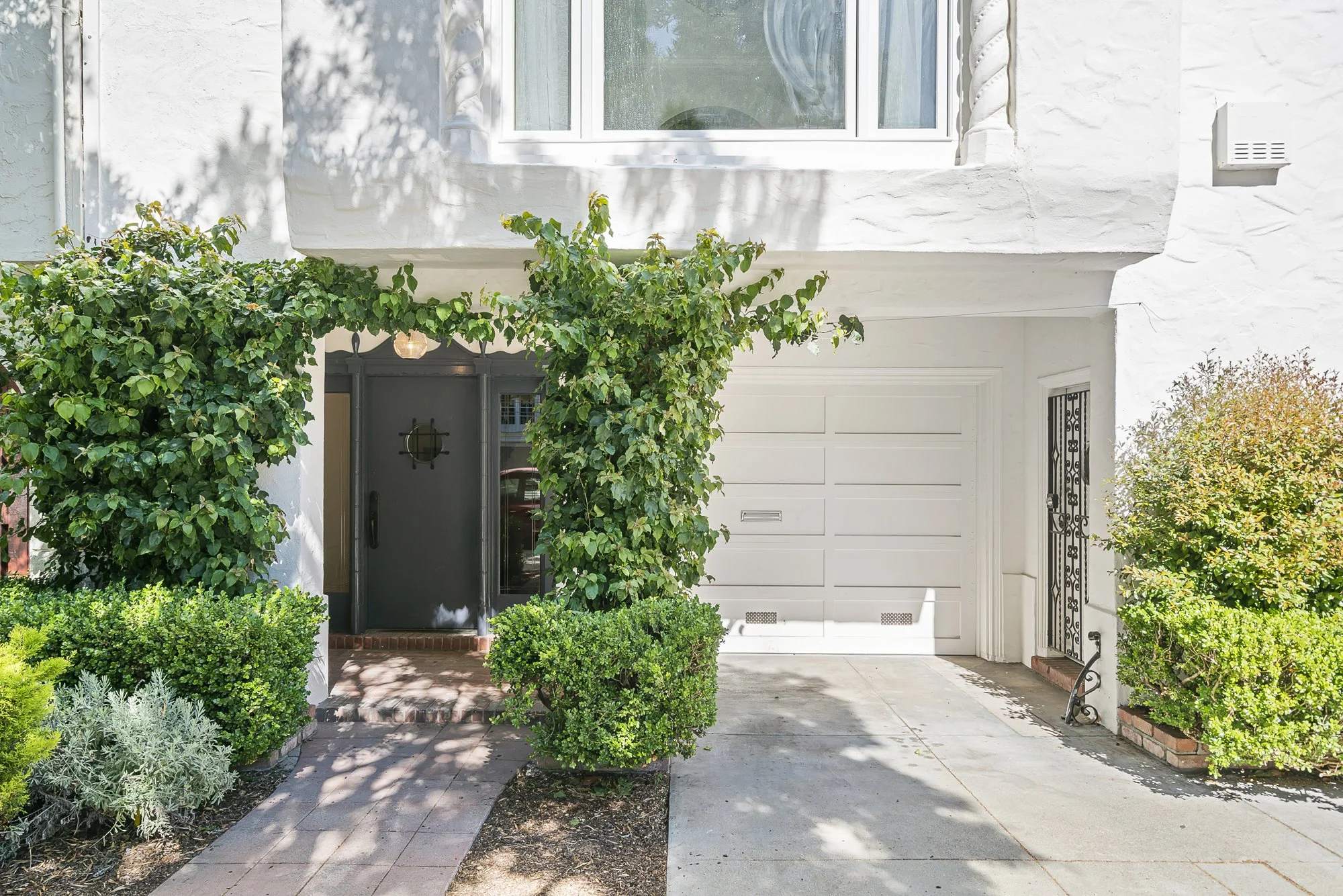 Front entry of a white house with a dark door framed by a climbing-vine arbor, landscaping, and a white garage door