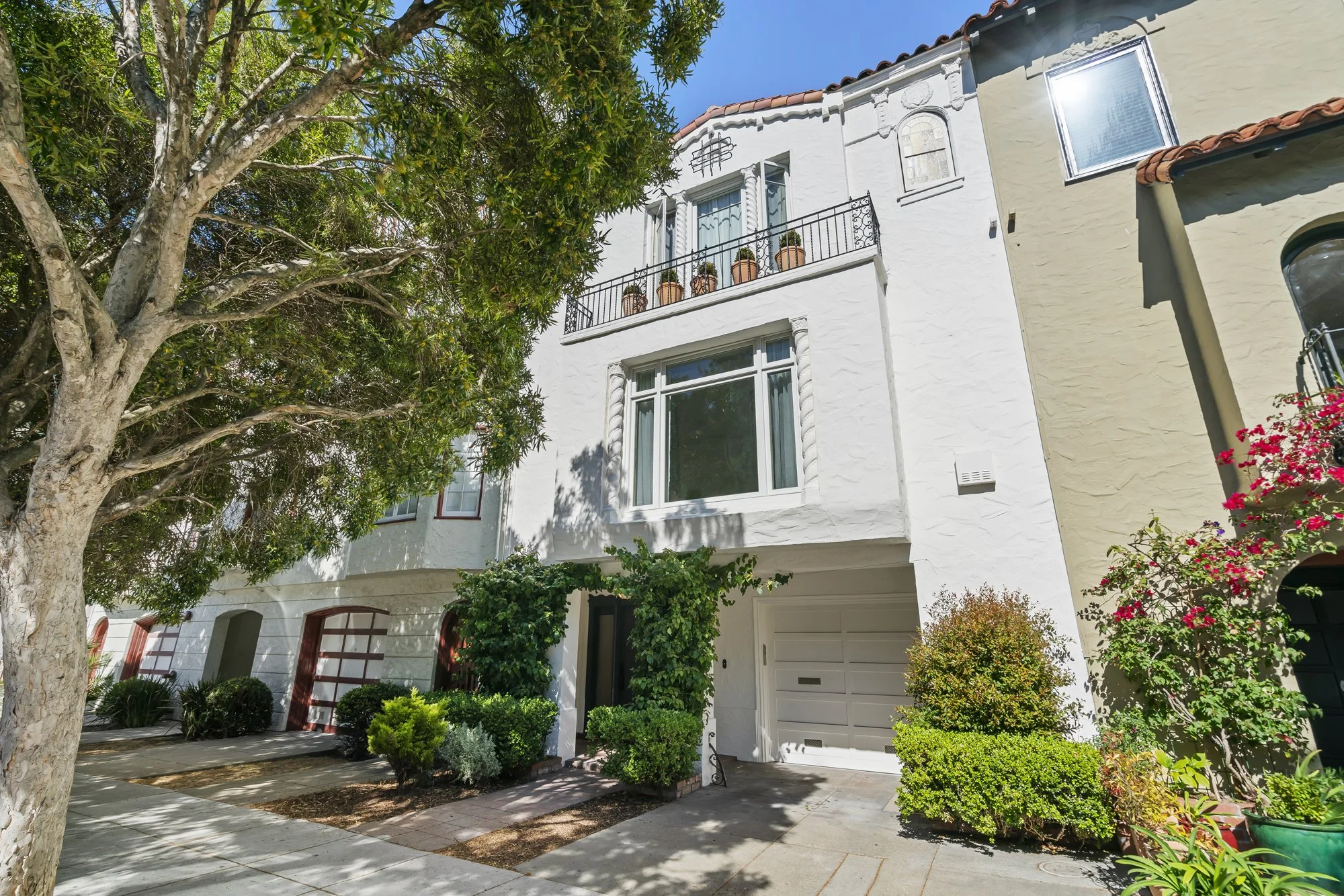 Street view of a white multi-level Mediterranean-style home with balconies, mature trees, and a landscaped front yard