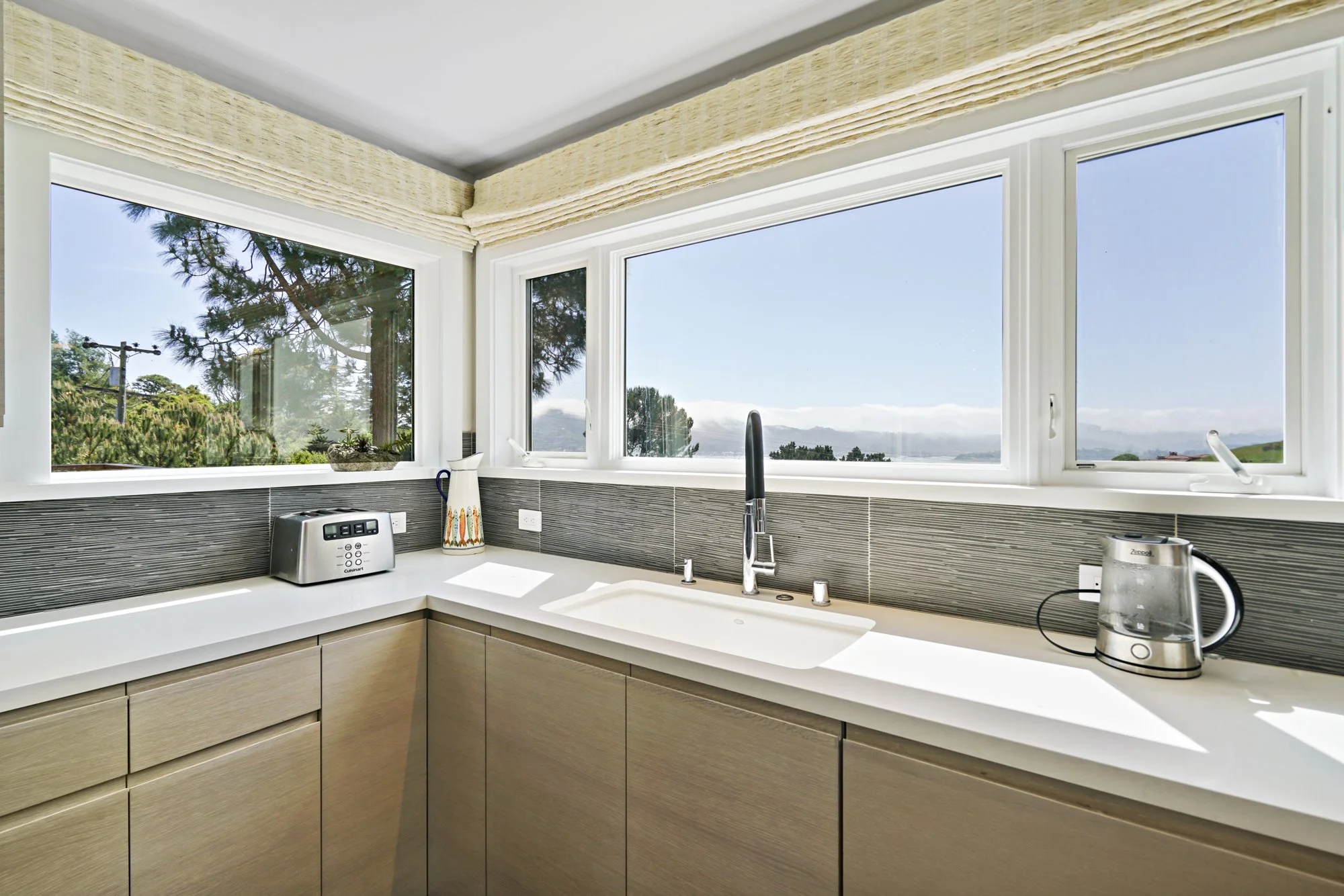 Kitchen corner with a stainless sink, gray tile backsplash, light wood cabinets, and wraparound windows with a bay view