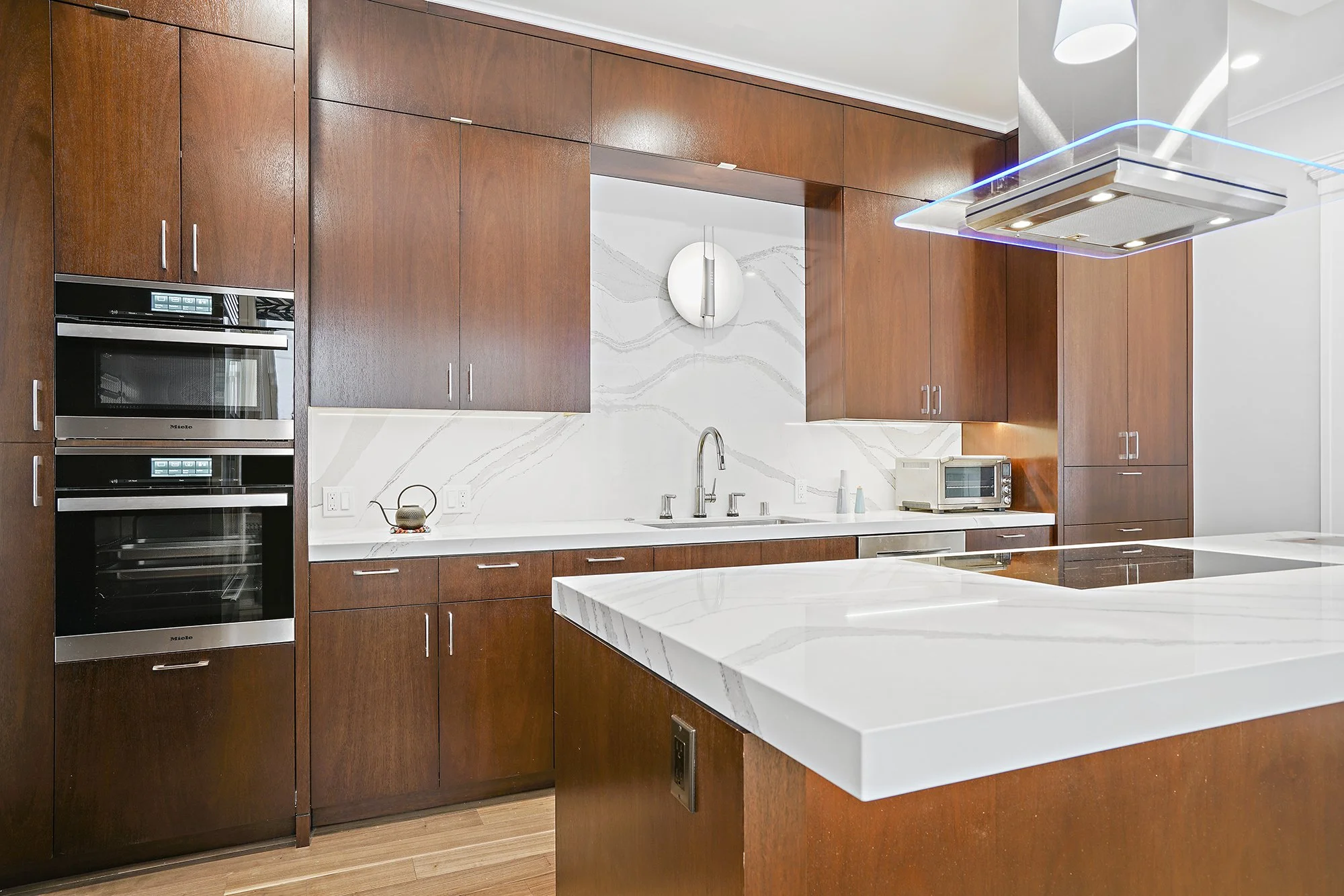 Wood kitchen with a marble waterfall island, marble backsplash, built-in stainless ovens, and a modern range hood