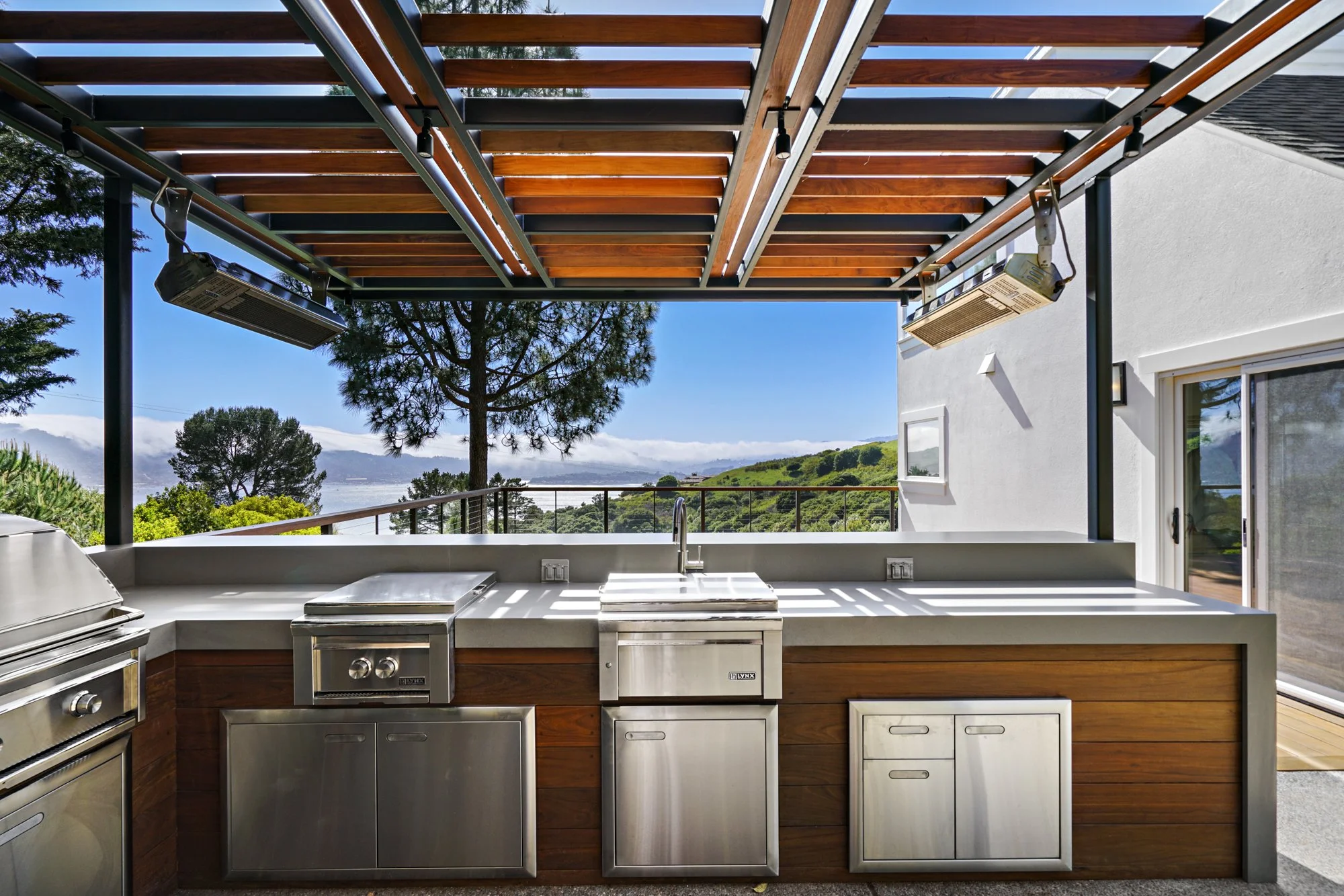 Covered outdoor kitchen with stainless appliances, an undermount sink, wood cabinetry, and a hillside bay view