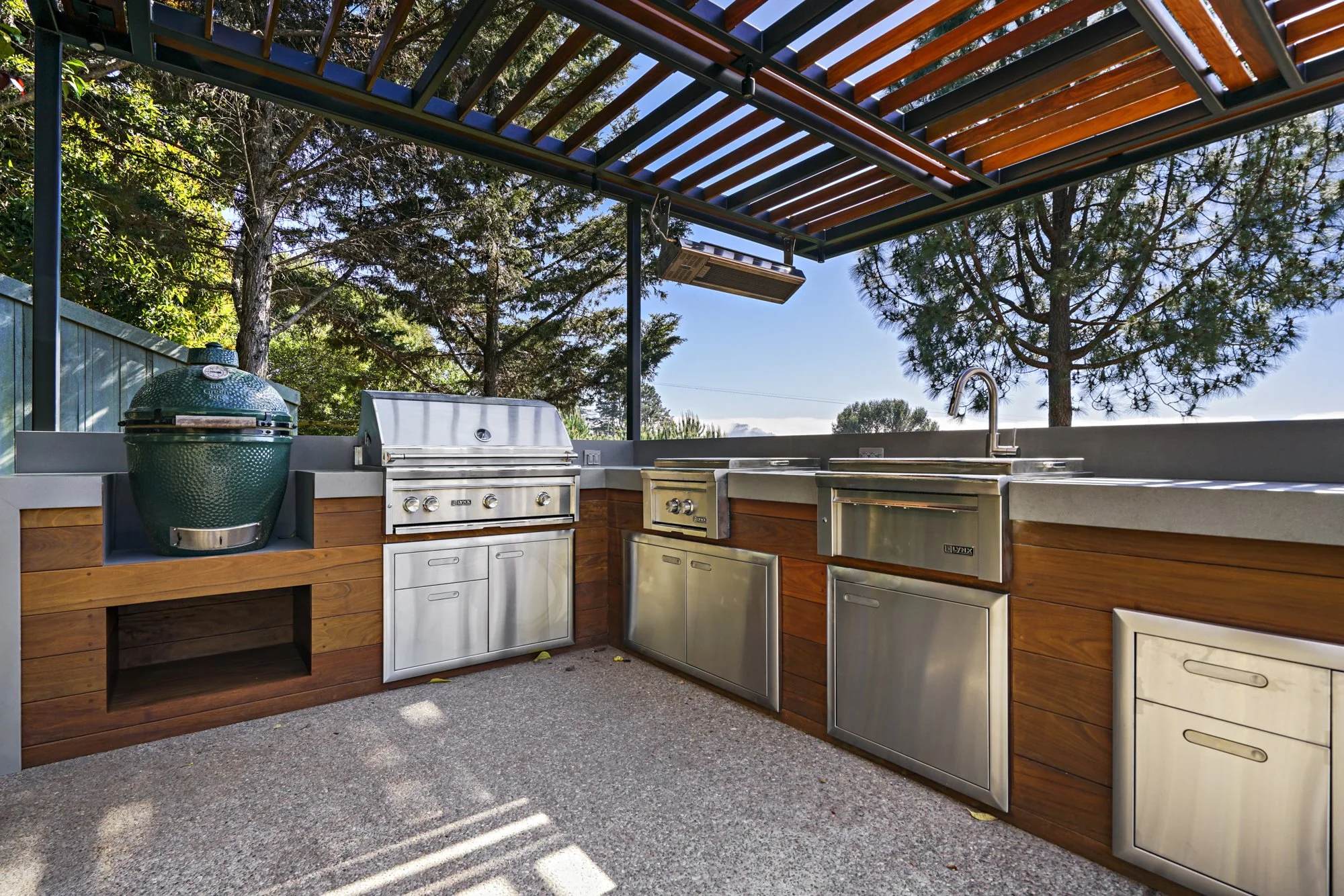Outdoor kitchen with a green ceramic egg smoker, stainless grill, wood cabinets, and a glass-railed deck overlooking the bay