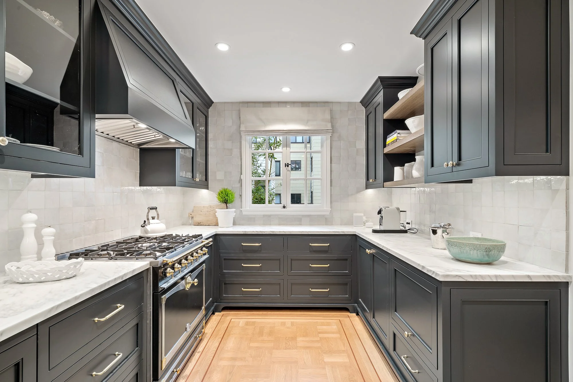 U-shaped gray kitchen with a black range hood, marble counters and backsplash, brass hardware, and herringbone floors