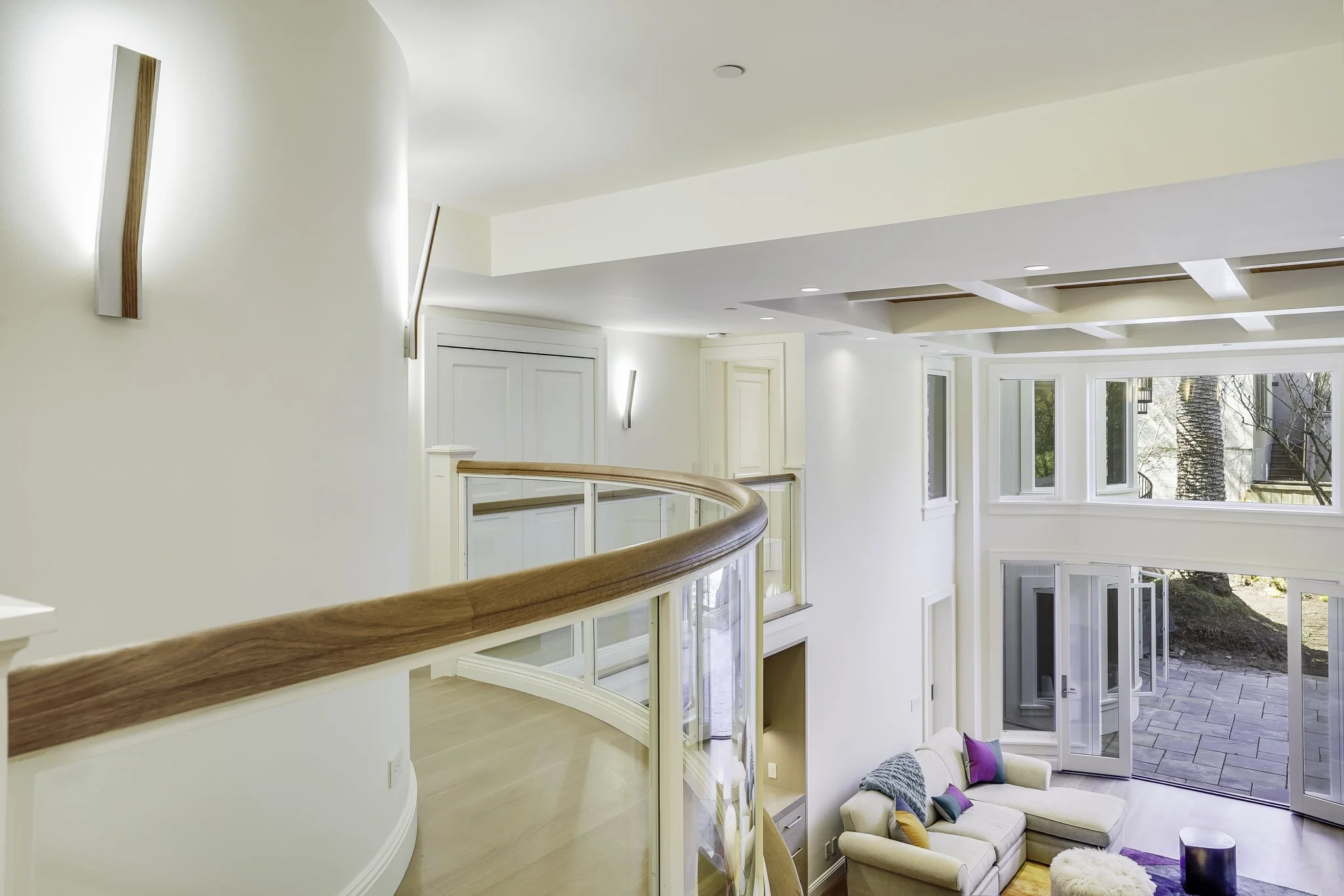 Upper-level walkway with a curved wood-and-glass railing overlooking the living room below, and a coffered ceiling