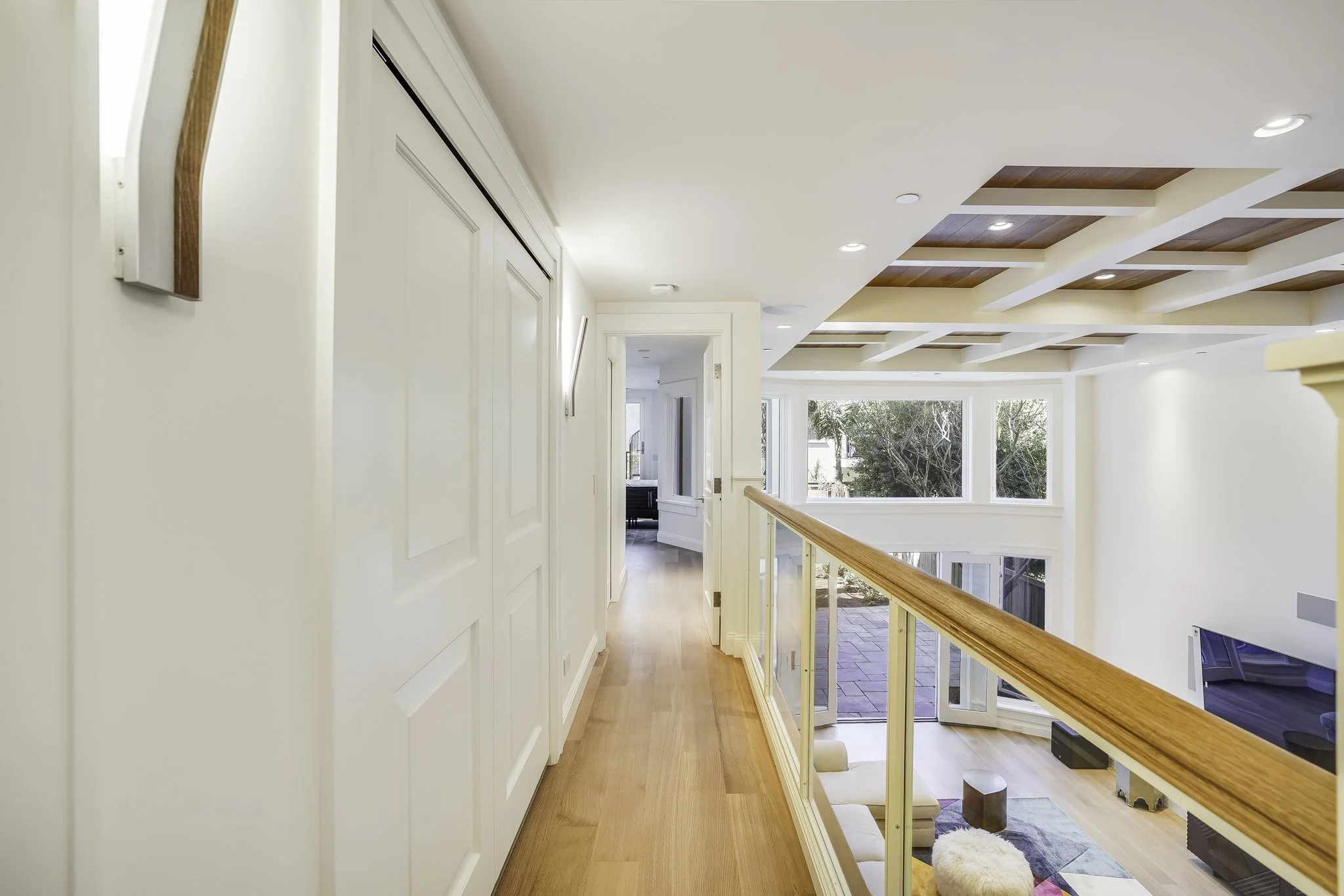 Hallway with a coffered wood ceiling, glass railing, white doors, and hardwood floors overlooking the atrium