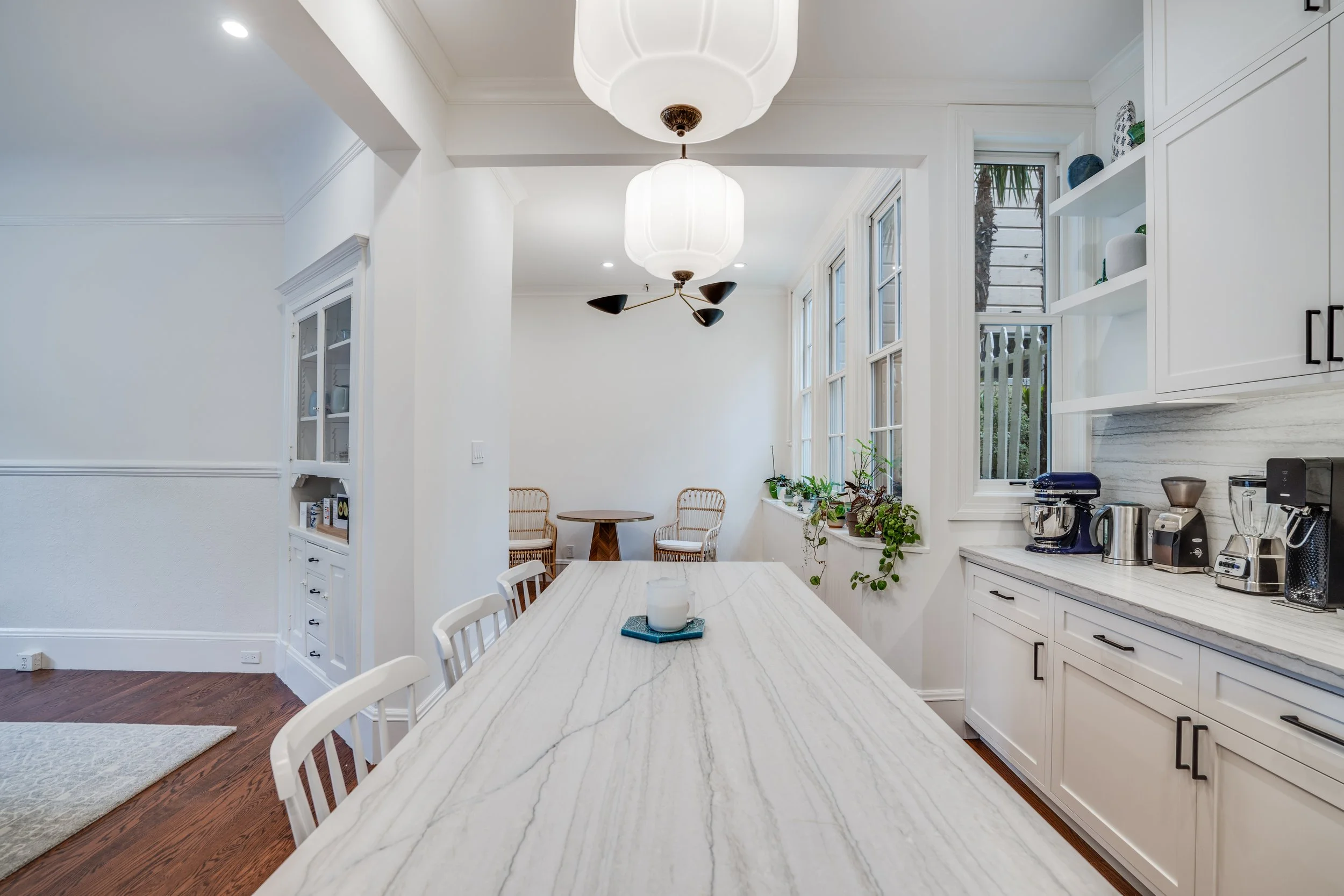 White kitchen and dining area with a long marble-look island, globe pendant lights, built-in cabinetry, and tall windows
