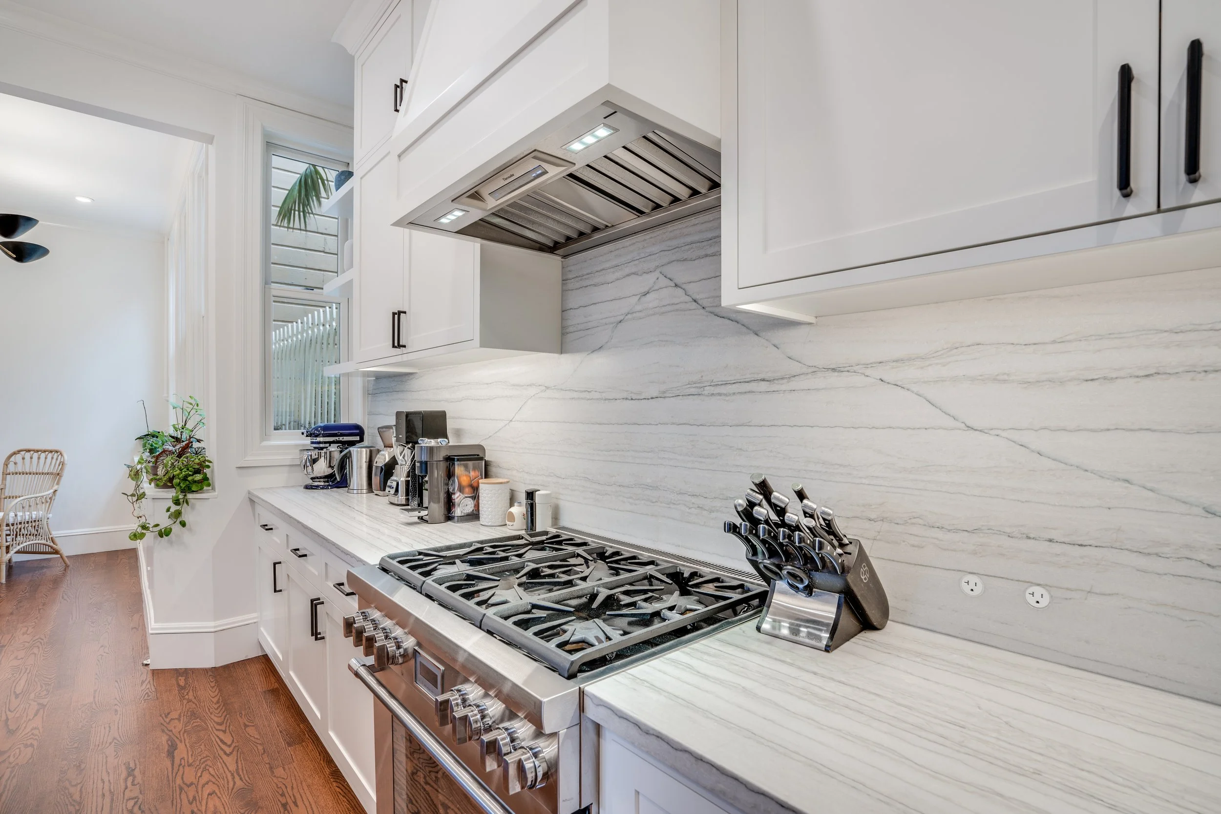 White kitchen with a stainless gas range, marble-look slab backsplash, matte-black hardware, and wood floors