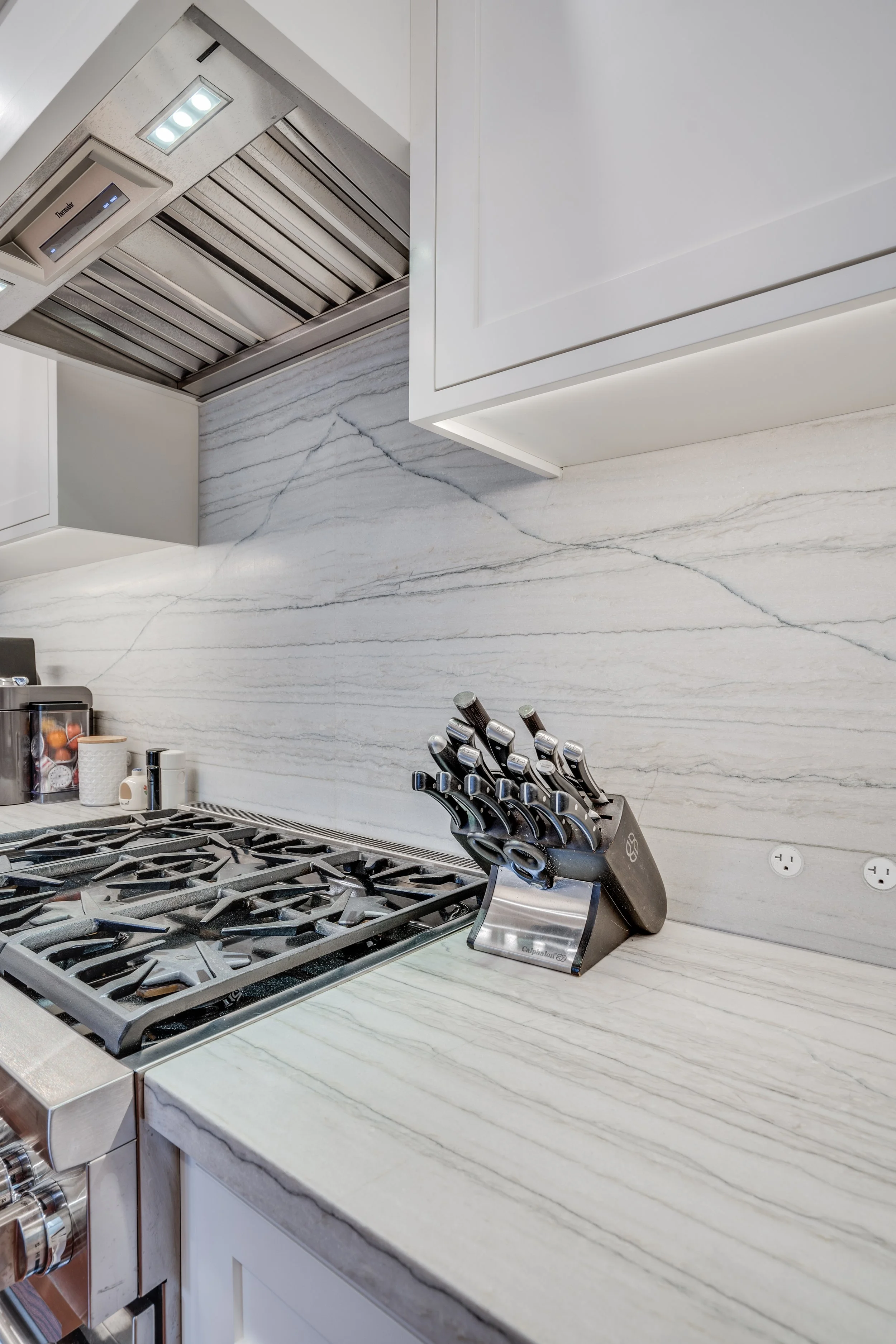 Close-up of a stainless gas cooktop with a marble-look slab backsplash and a black knife block