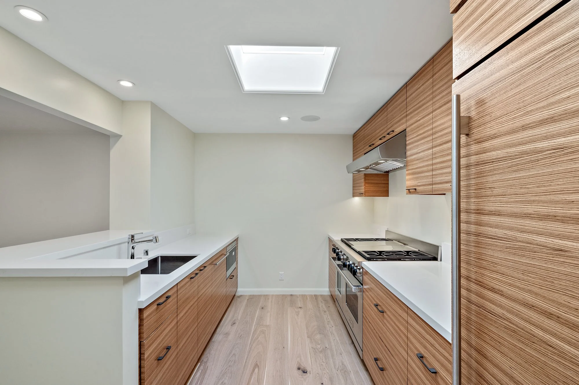 Galley kitchen with wood-grain cabinetry, stainless cooktop, white counters and a pass-through opening, light wood floors, and a skylight
