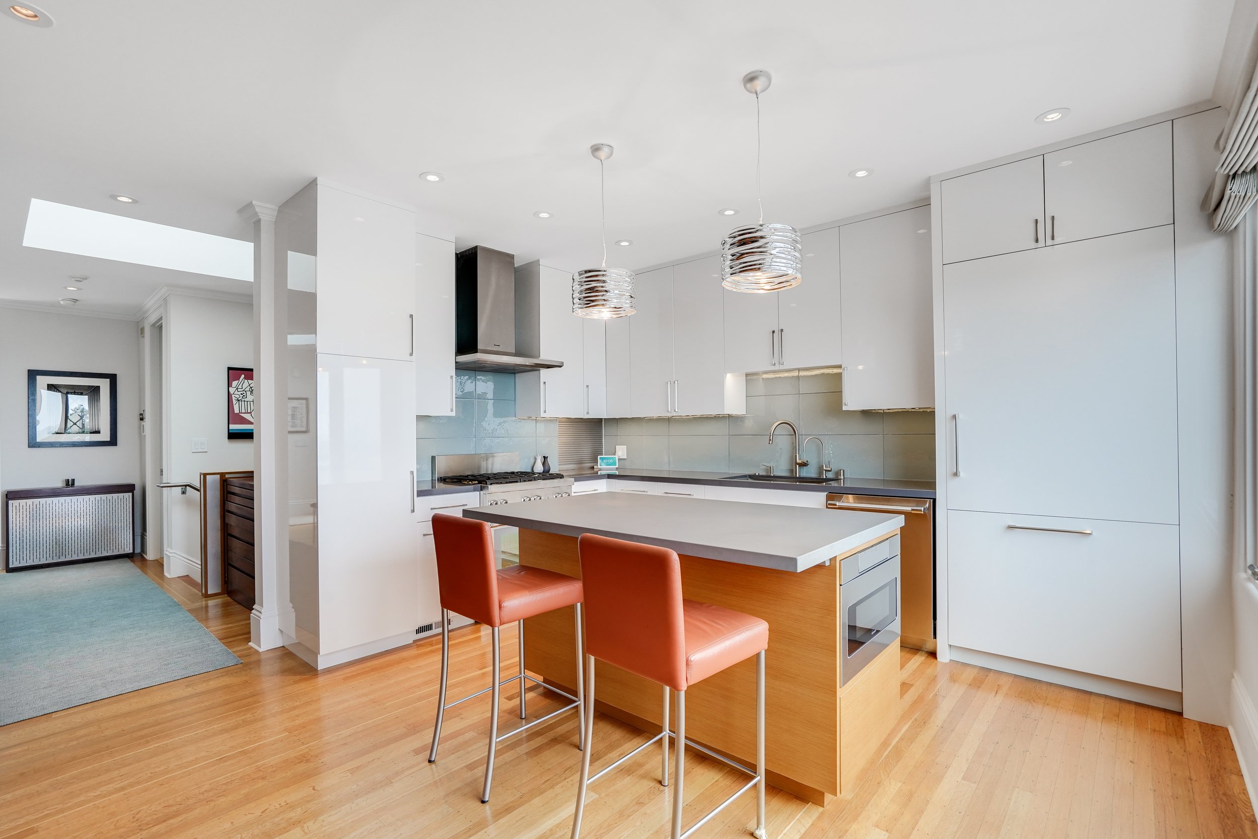 Modern white kitchen with a wood-topped island, rust leather bar stools, stainless appliances, drum pendant lights, and light wood floors