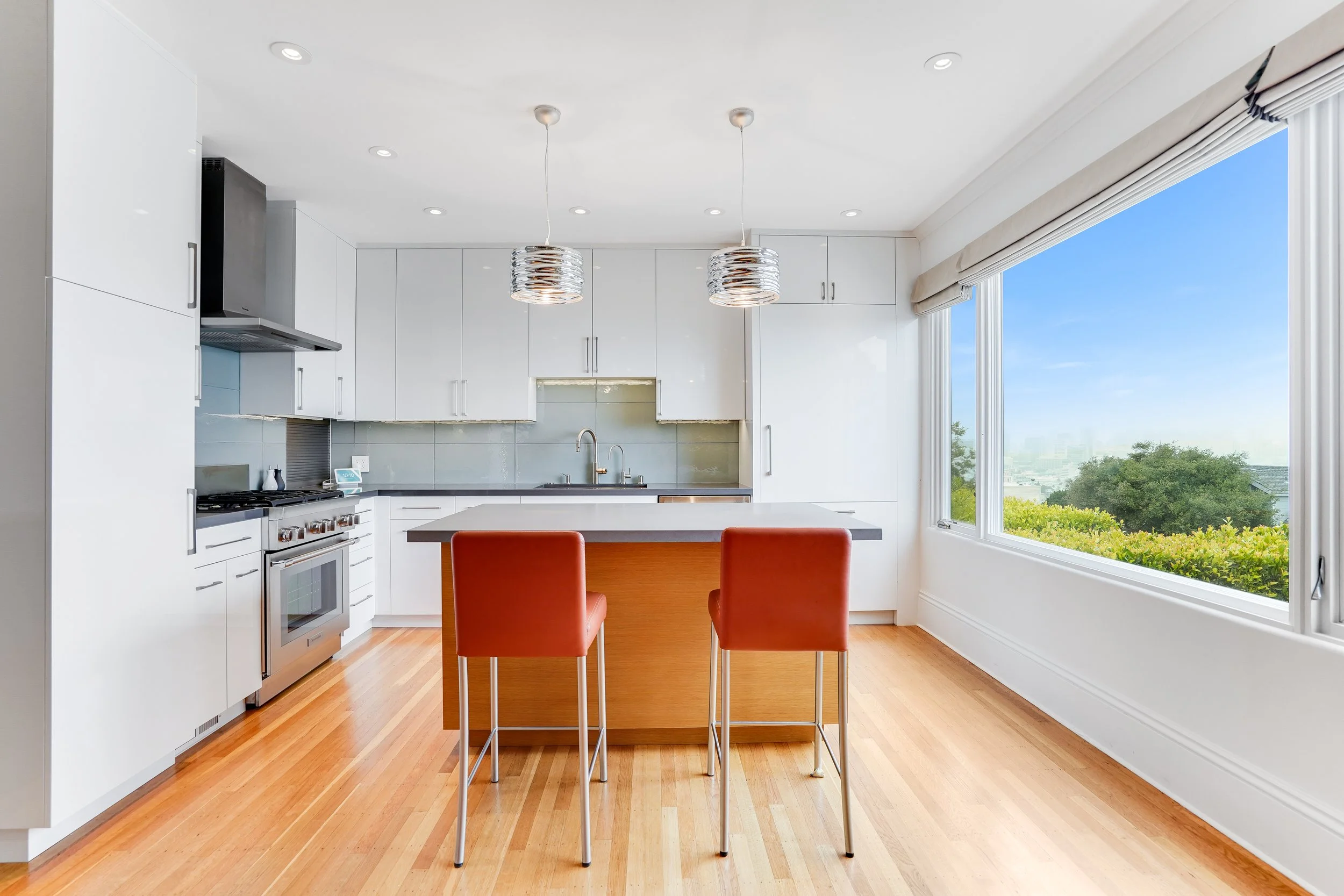 White kitchen with a wood island, rust leather stools, stainless range and hood, and corner windows with a green hillside view