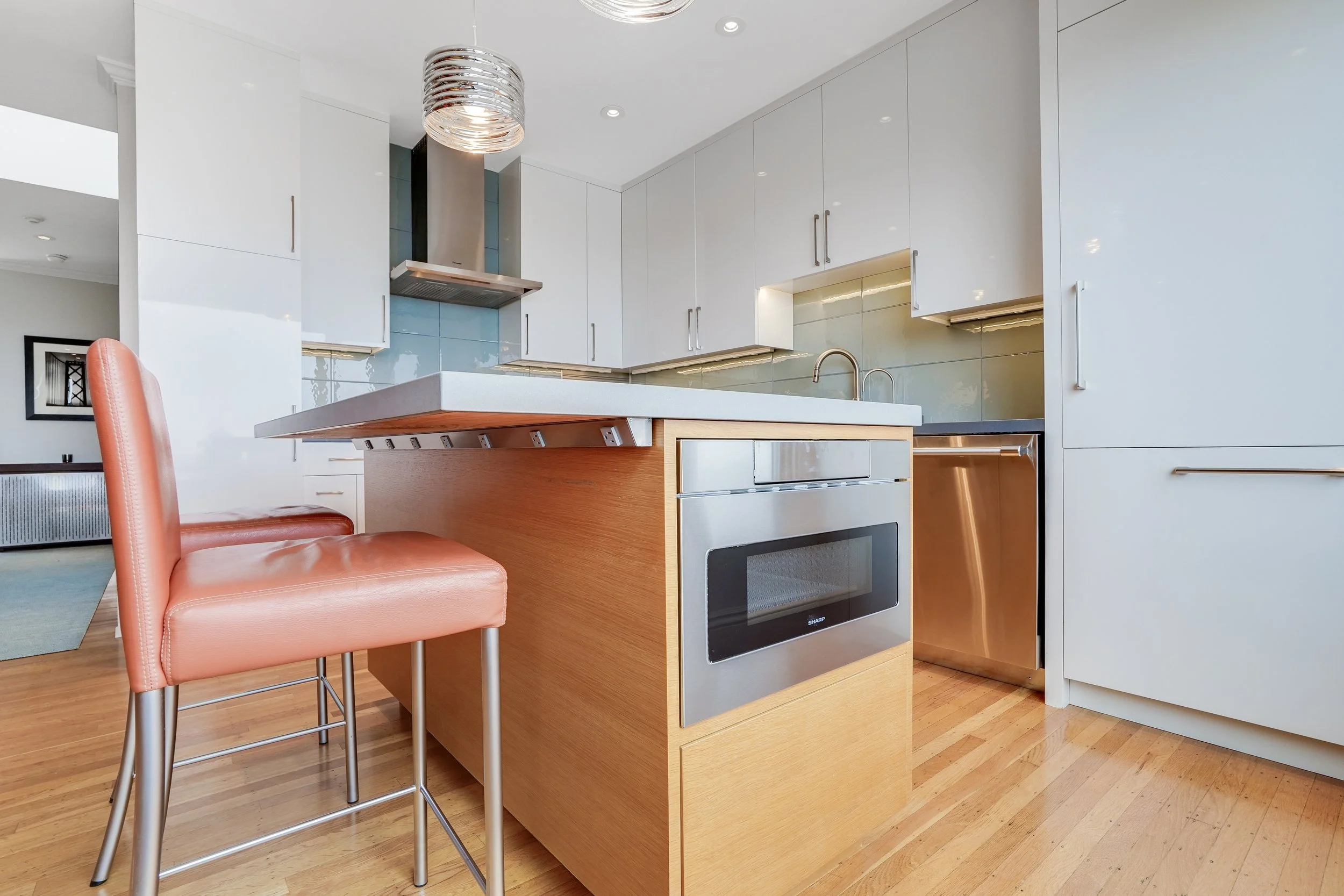 White kitchen with a wood-paneled island, rust leather bar stool, glass-tile backsplash, and stainless built-in microwave