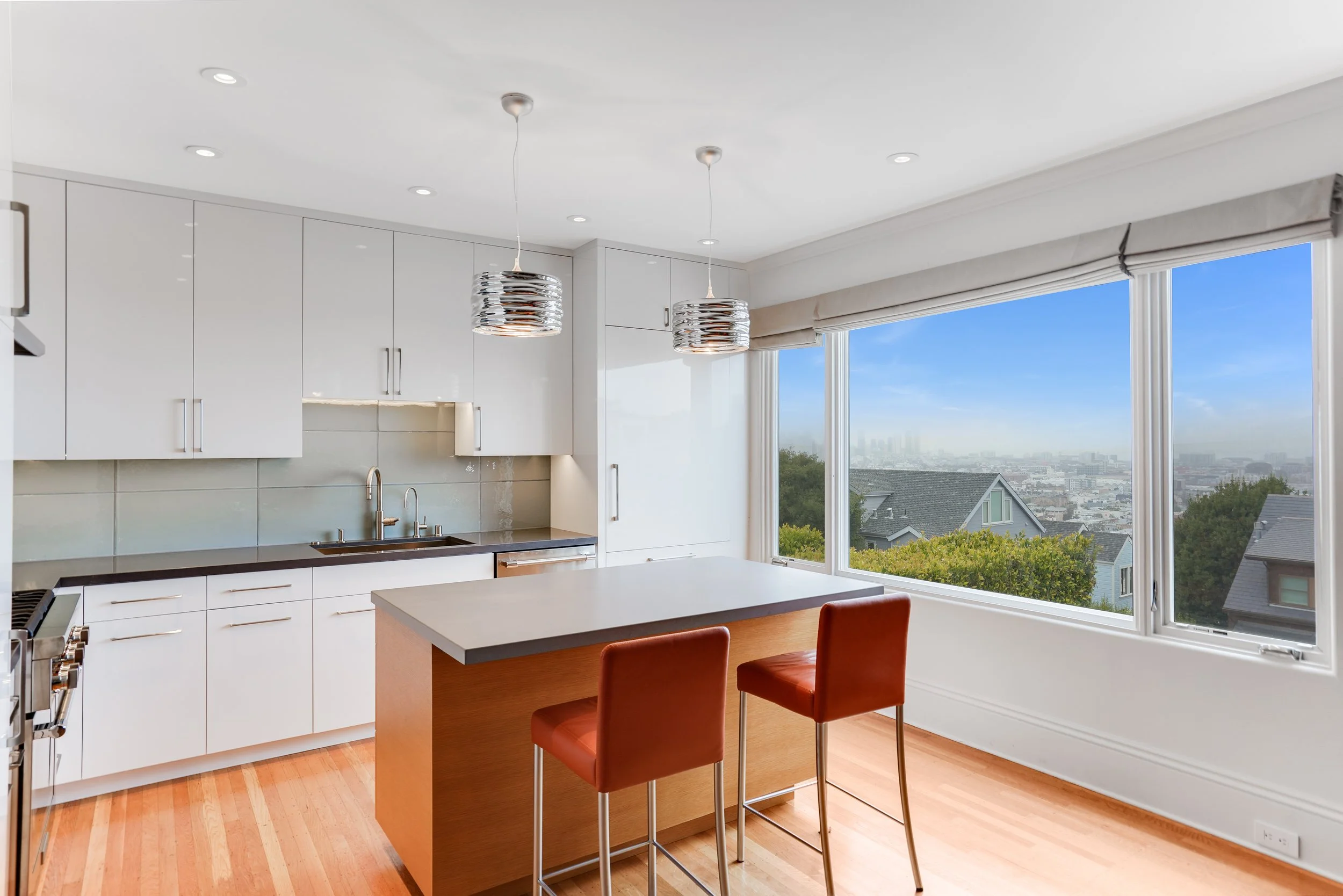 White kitchen with a wood-topped island, rust leather stools, and large corner windows with a panoramic city view